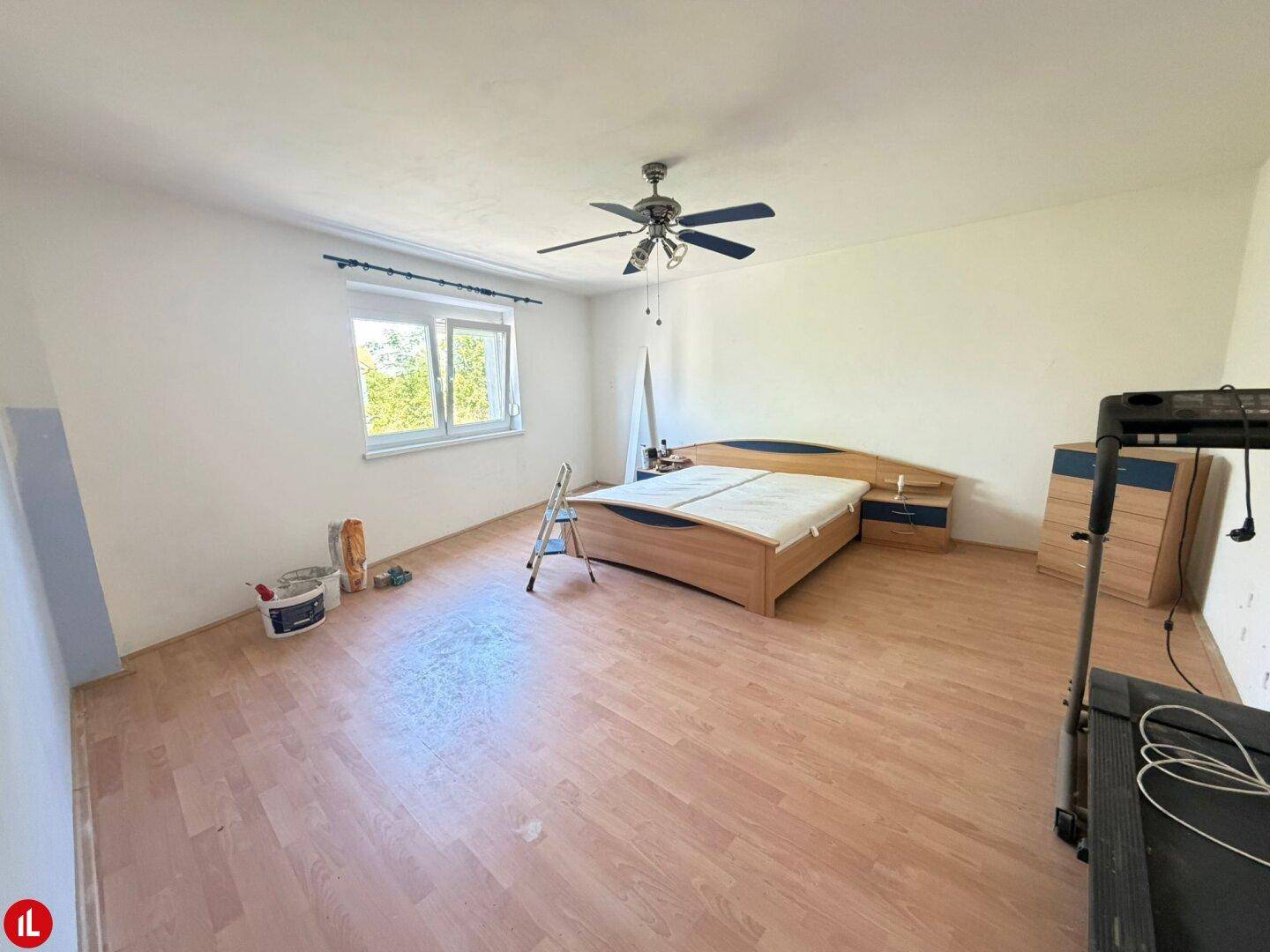 Schlafzimmer mit Bett, Deckenventilator, Laminatboden und Fenster, bereit zur Renovierung.