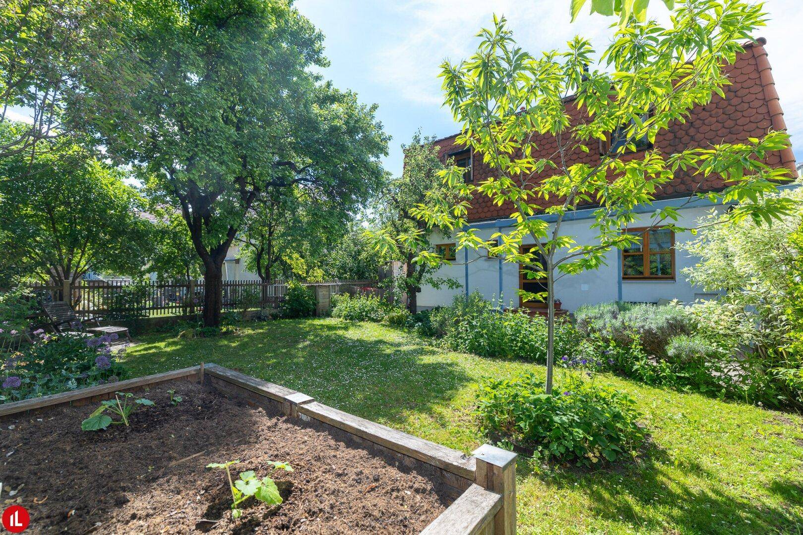 Gepflegter Garten mit Rasenfläche, Hochbeet und altem Baumbestand vor einem charmanten Haus mit Ziegeldach.