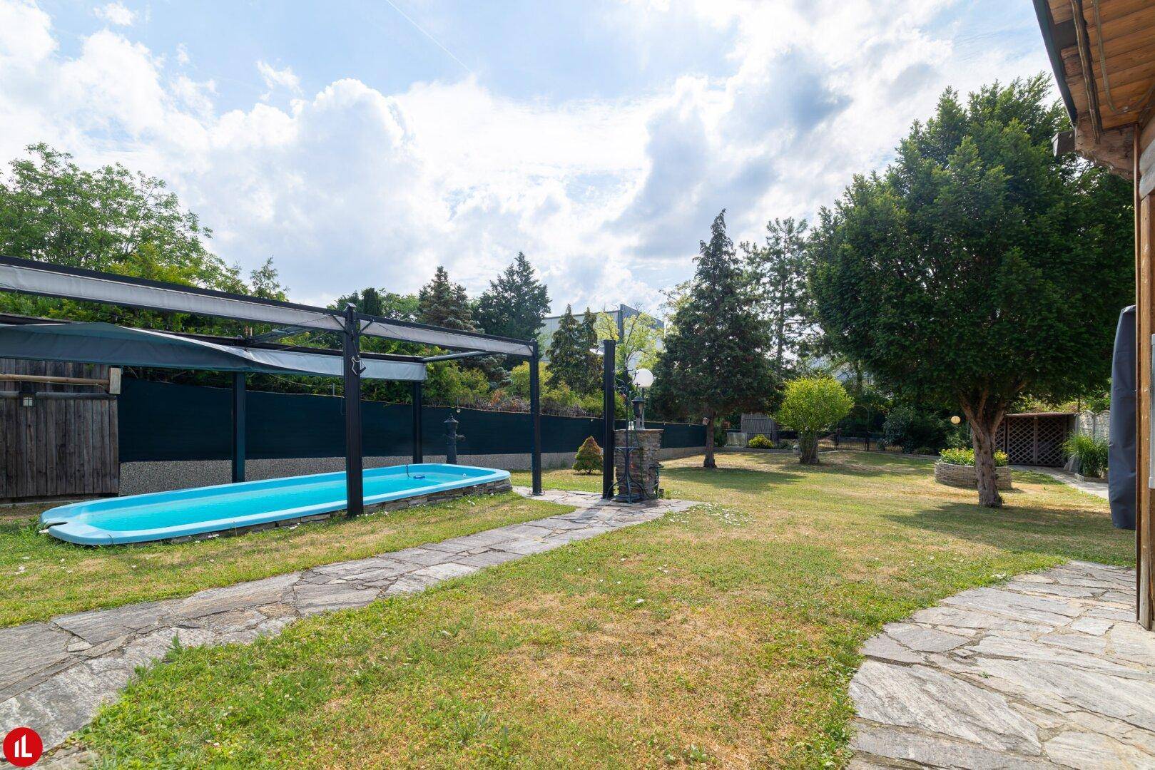 Weitläufiger Garten mit einem eingelassenen Pool, Steinwegen und üppiger Vegetation unter bewölktem Himmel.