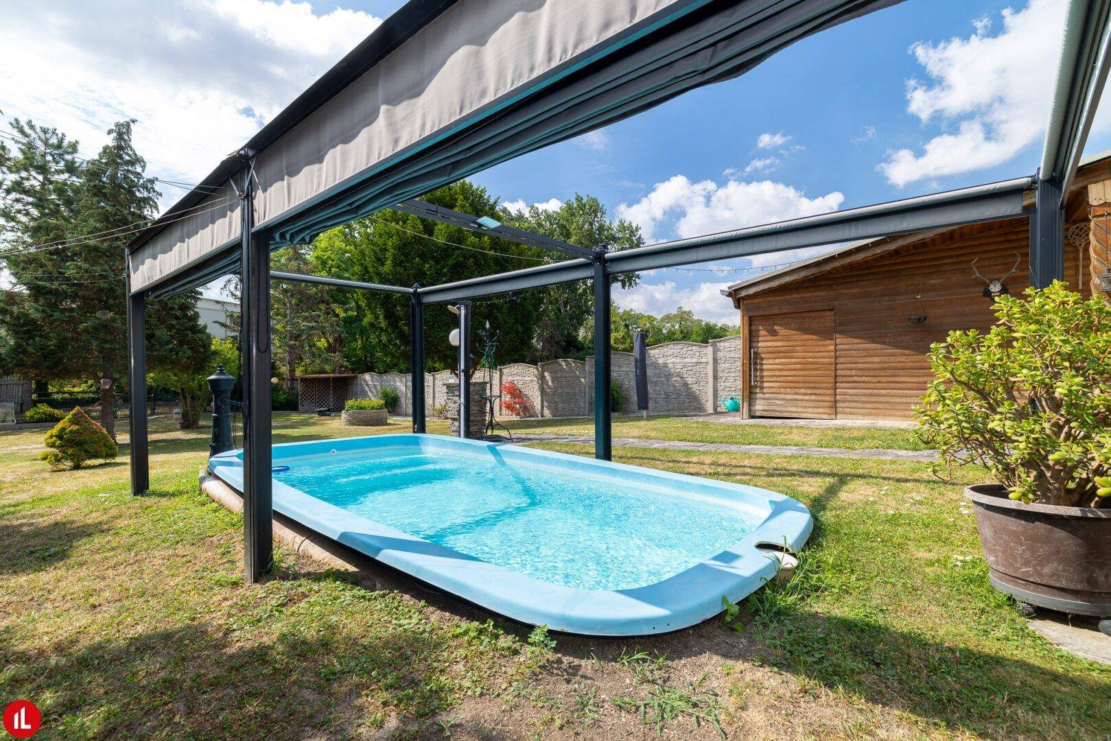 Blauer Pool im Garten unter einer Überdachung, umgeben von Rasen und Pflanzen an einem sonnigen Tag.