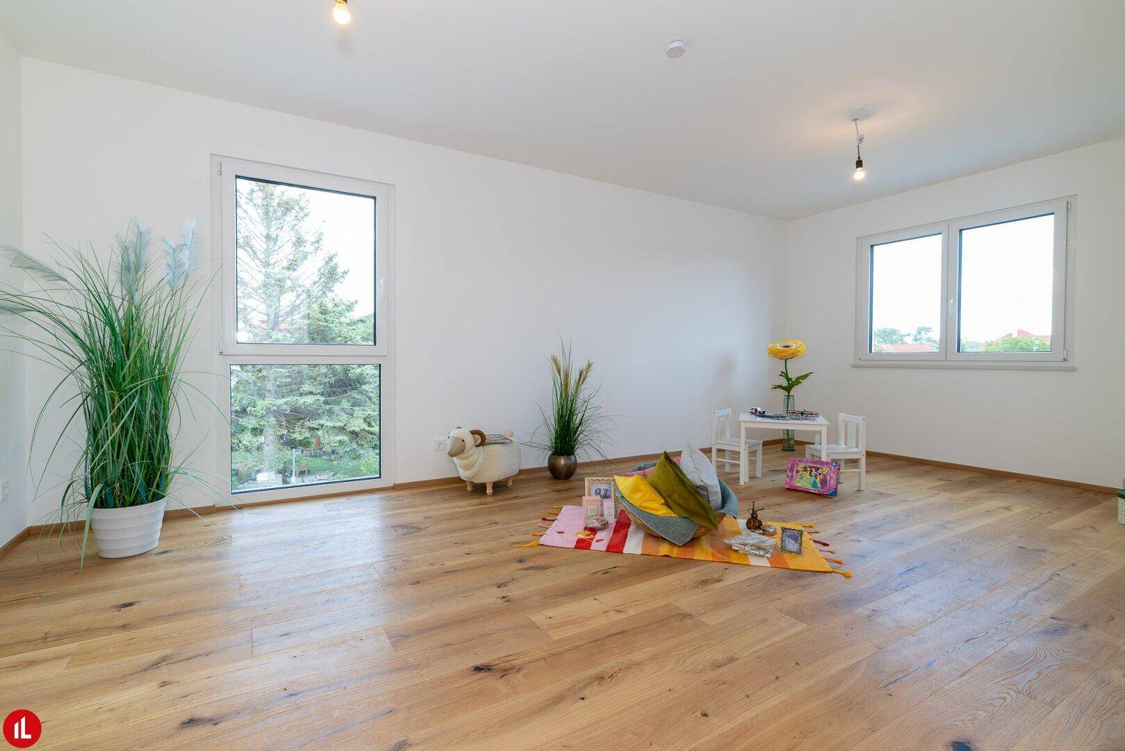 Helles Zimmer mit Holzboden und zwei großen Fenstern mit Blick ins Grüne.