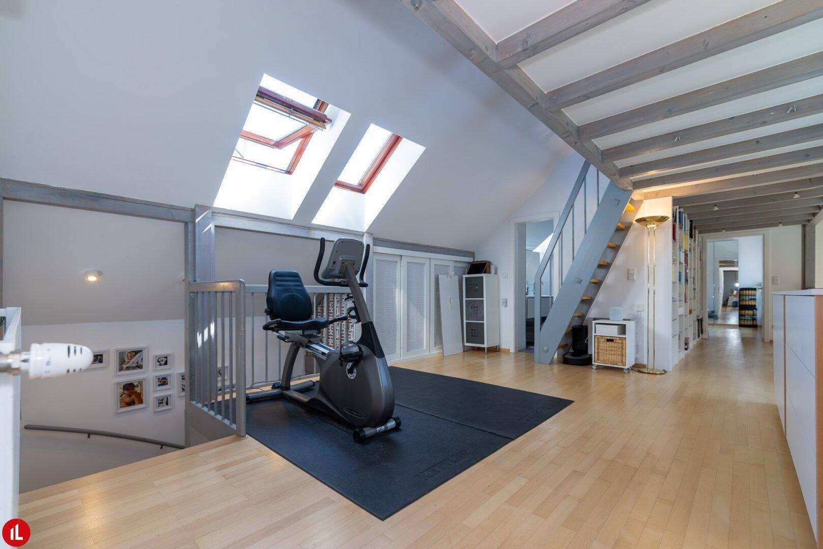 Offener Bereich im Dachgeschoss mit Fitnessgerät, Dachfenstern und Holzbalkendecke.