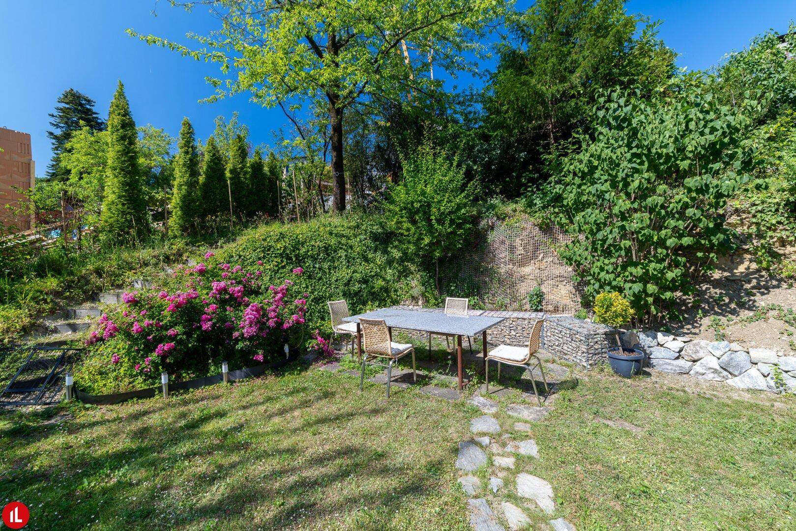 Sonniger Gartenbereich mit Rasenfläche, blühenden Sträuchern und Esstisch im Freien.