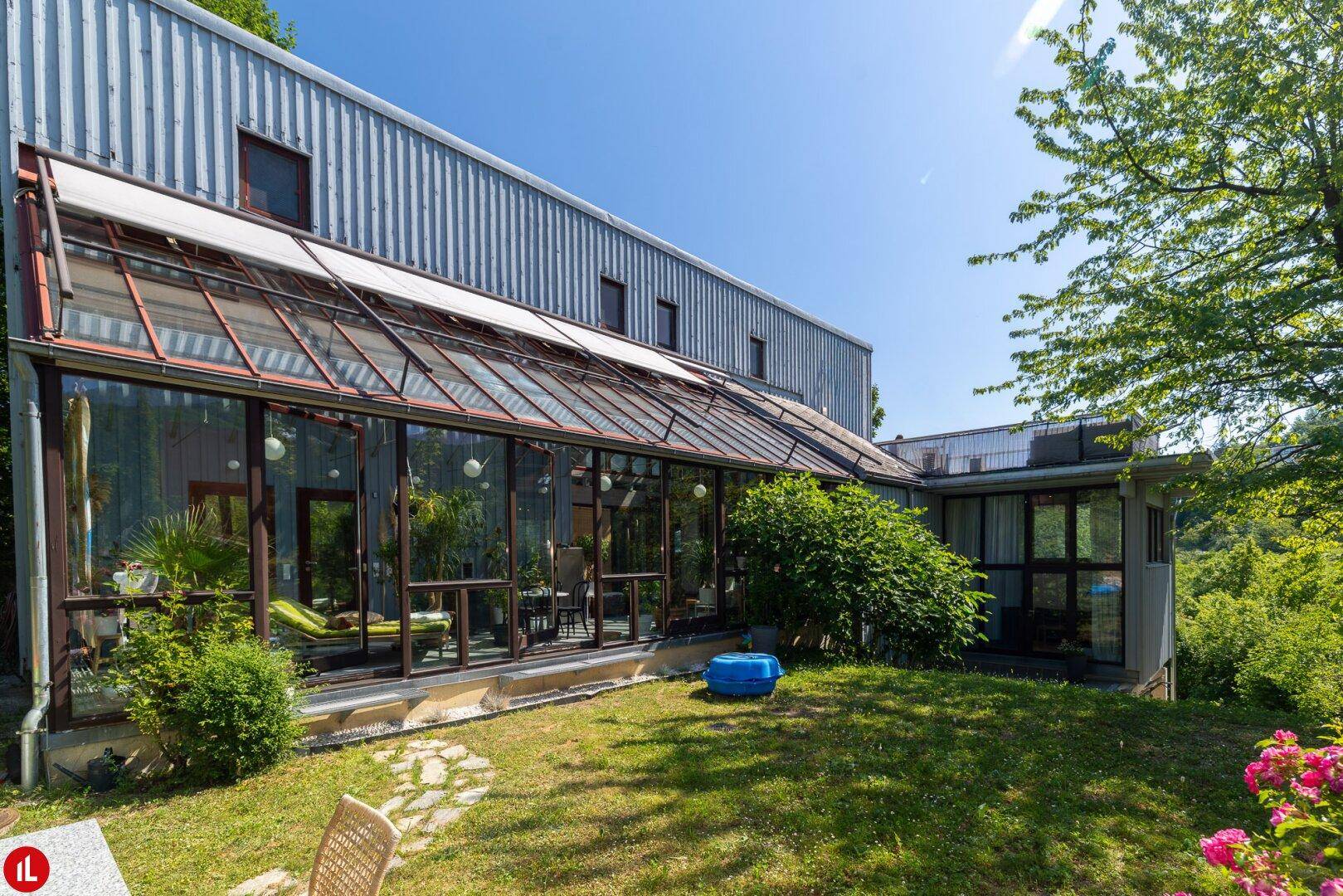 Modernes Gebäude mit Glasanbau, umgeben von viel Grün und sonnigem Garten.