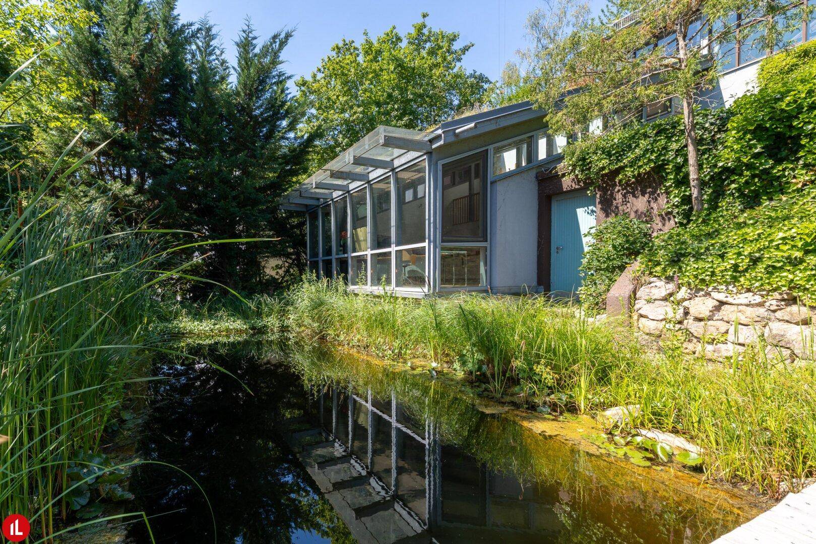 Idyllischer Garten mit Teich, Holzsteg und üppiger Vegetation rund um das Haus.