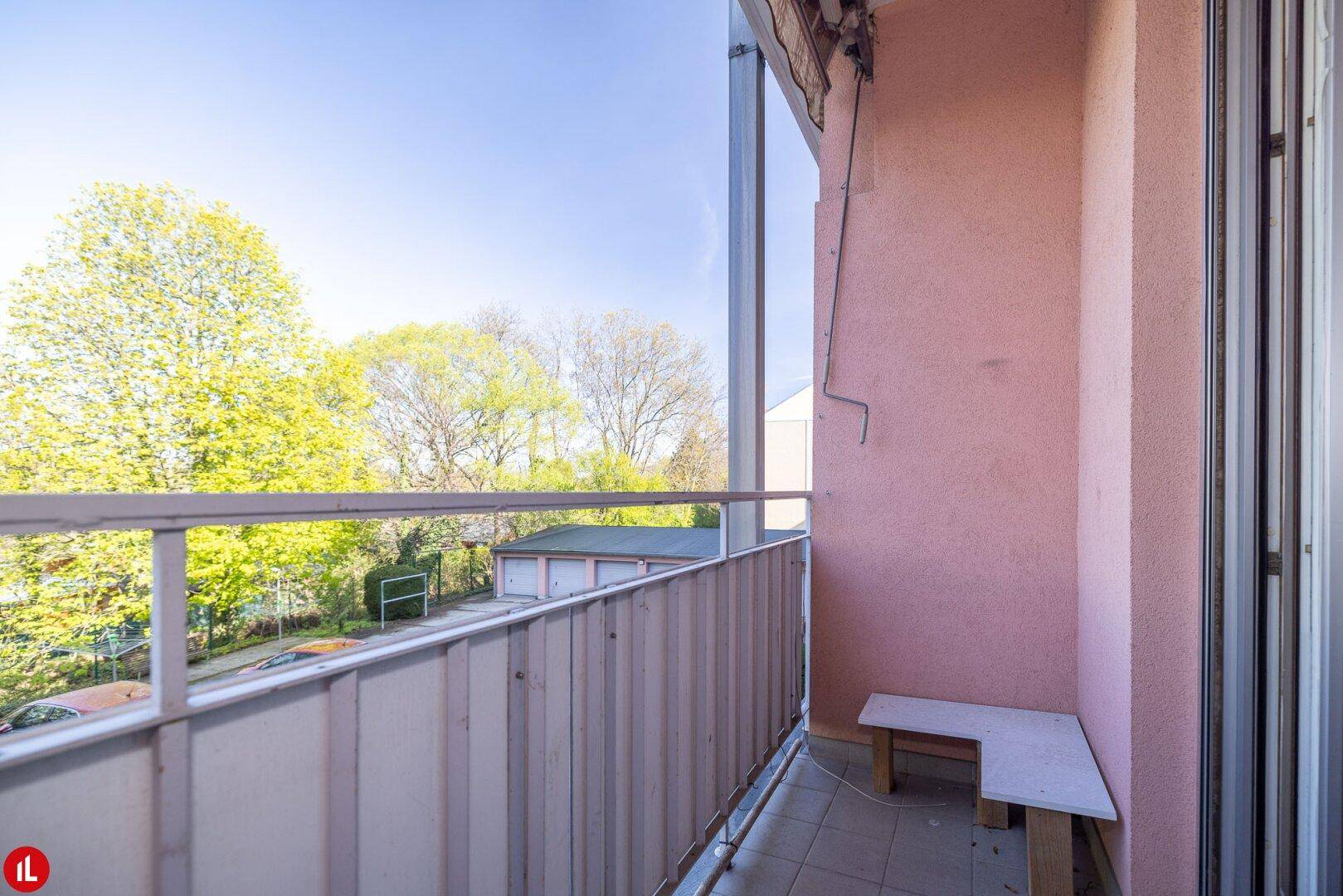Leerer Balkon mit rosa Fassade und Blick ins Grüne mit Bäumen und Garagen.