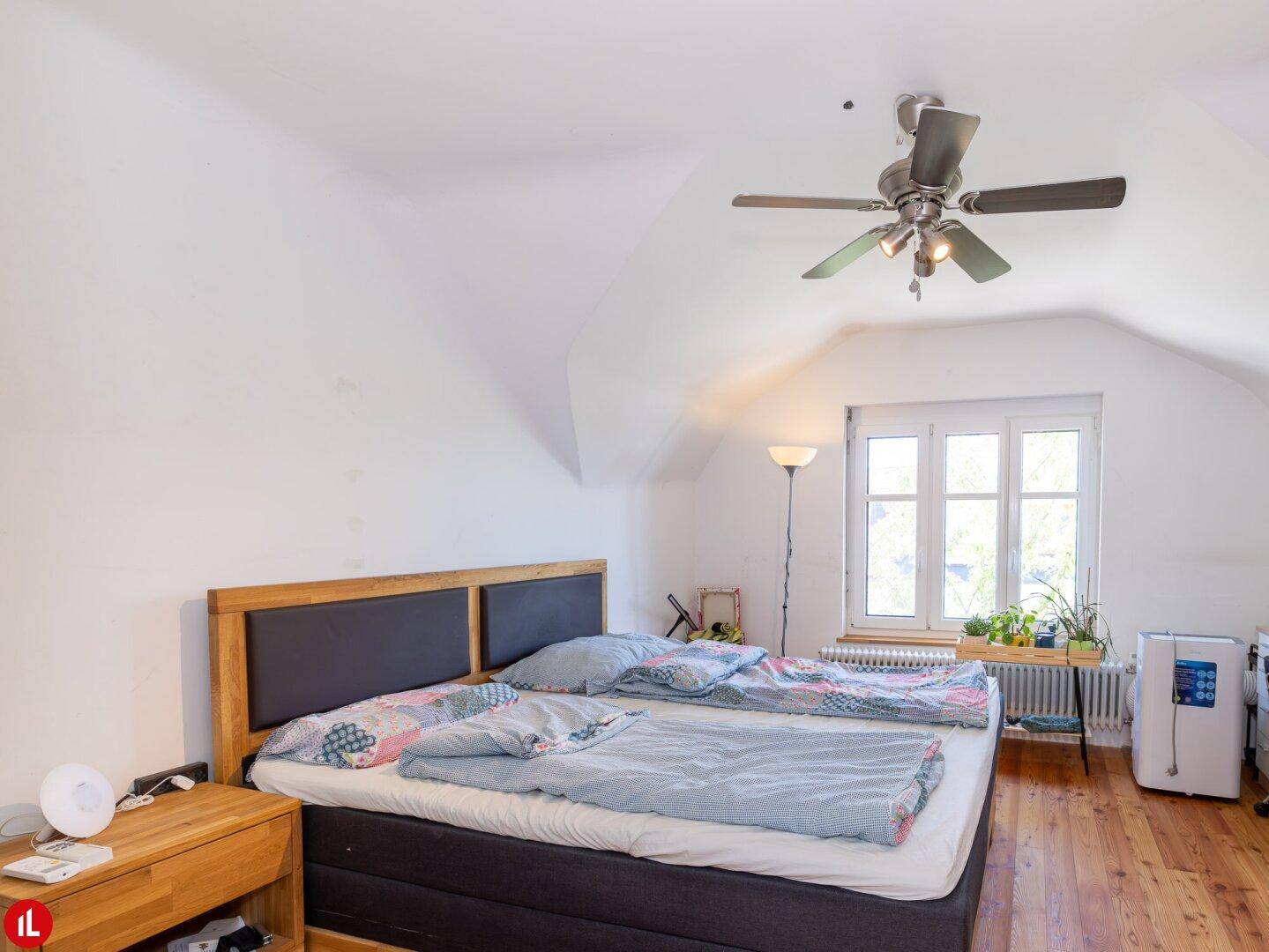 Schlafzimmer im Dachgeschoss mit Doppelbett, Holzmöbeln, Deckenventilator und schrägen Wänden.