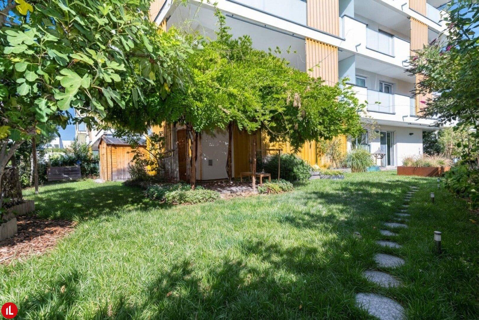 Sonniger Gartenbereich mit Rasen, Bäumen und einem kleinen Holzschuppen neben dem modernen Gebäude.