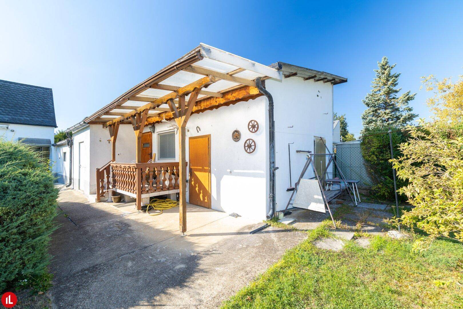 Überdachte Terrasse mit Holzgeländer und Zugang zum Nebengebäude, umgeben von Grün.