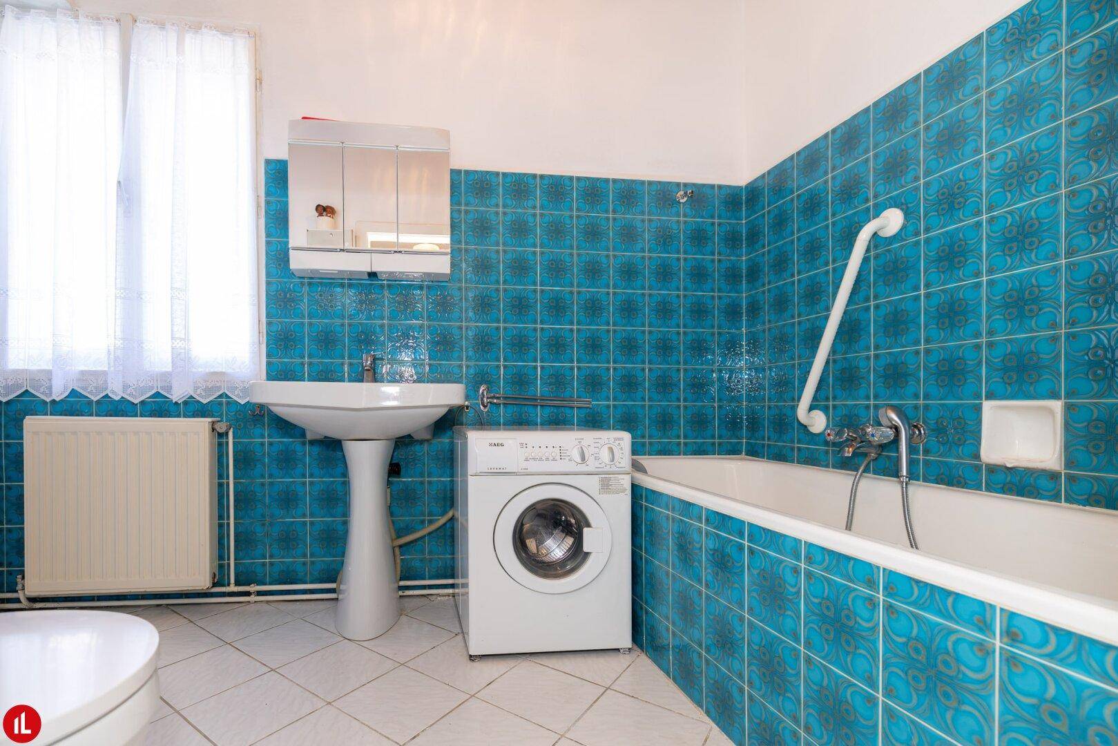 Badezimmer im Originalzustand mit blauen Fliesen, Badewanne und Waschmaschine.