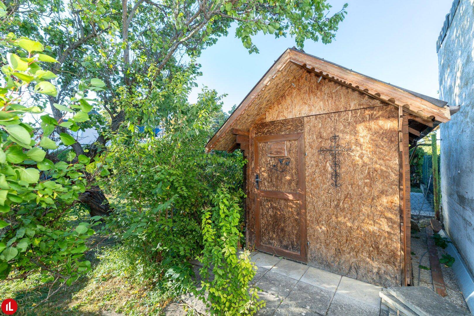 Rustikaler Gartenschuppen aus Holz im Grünen, umgeben von Bäumen und Sträuchern.