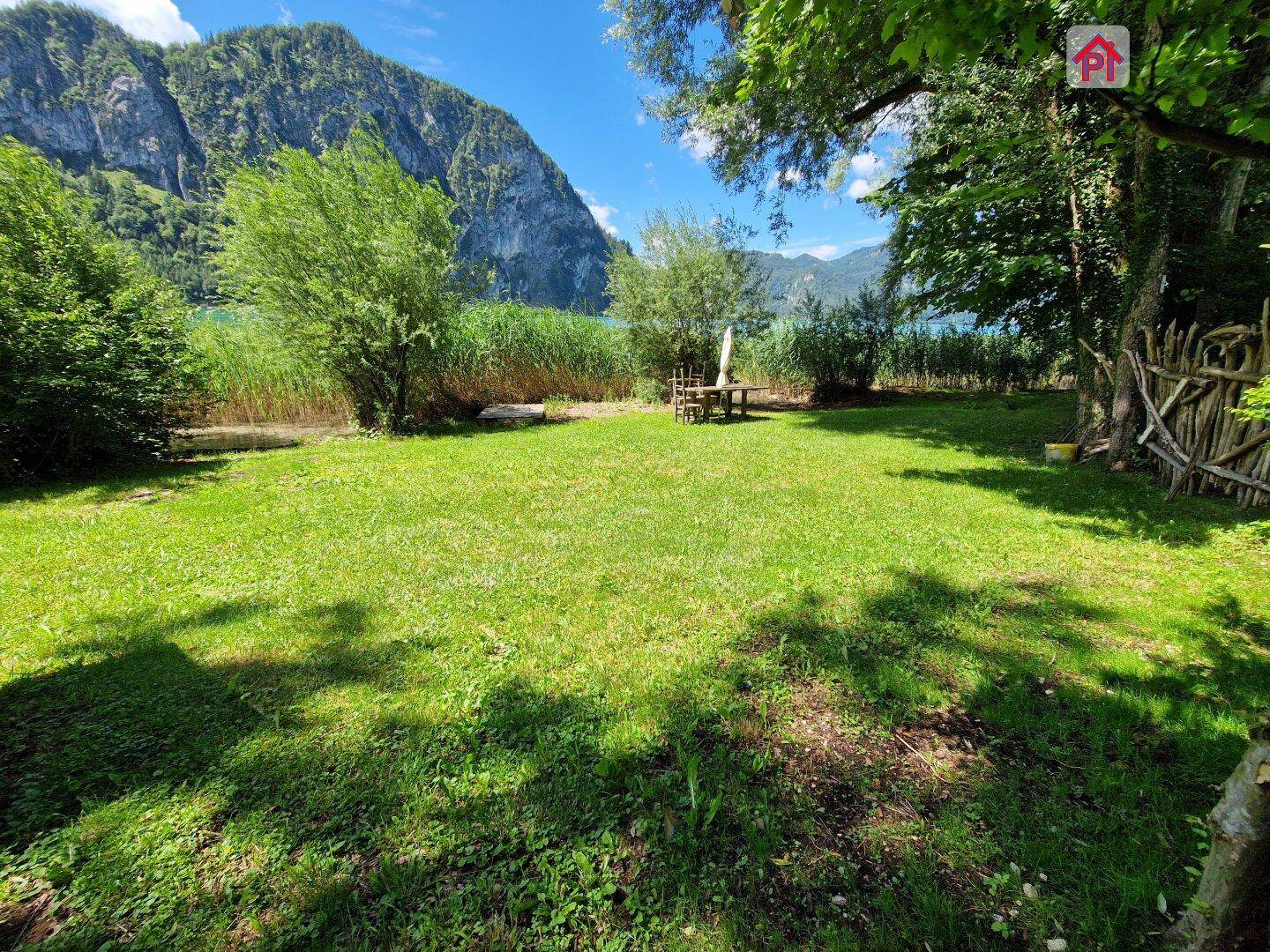 Weitläufige Rasenfläche im Garten mit Seezugang und umrahmt von Bäumen und Bergen.