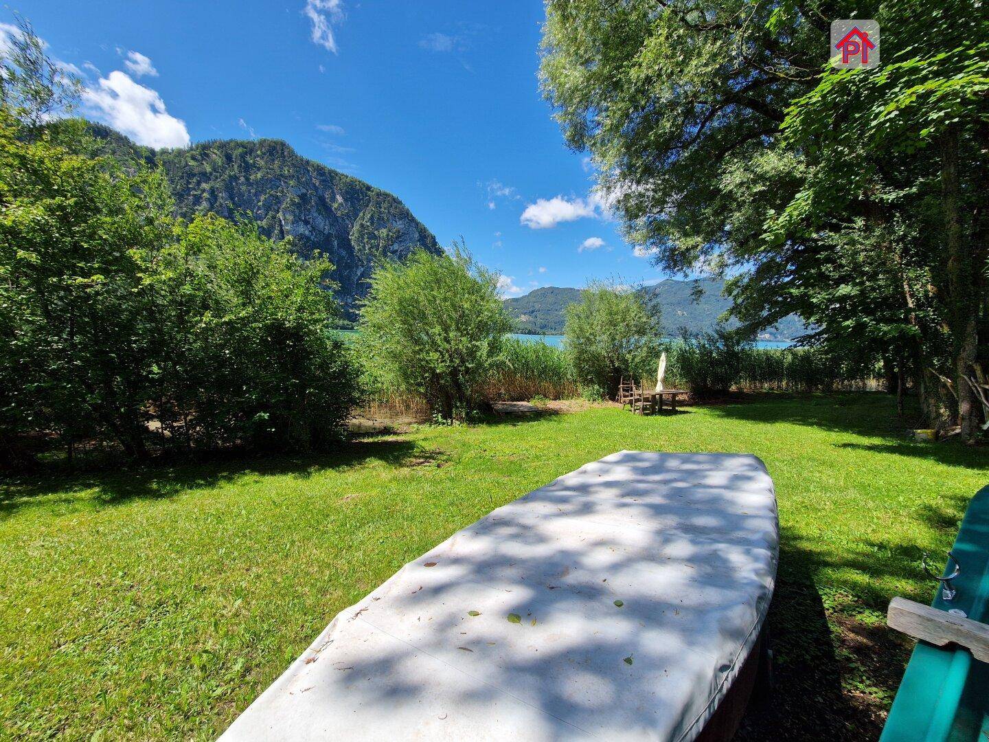 Sonniger Gartenbereich mit Rasenfläche, abgedecktem Boot und weitem Blick auf den See.
