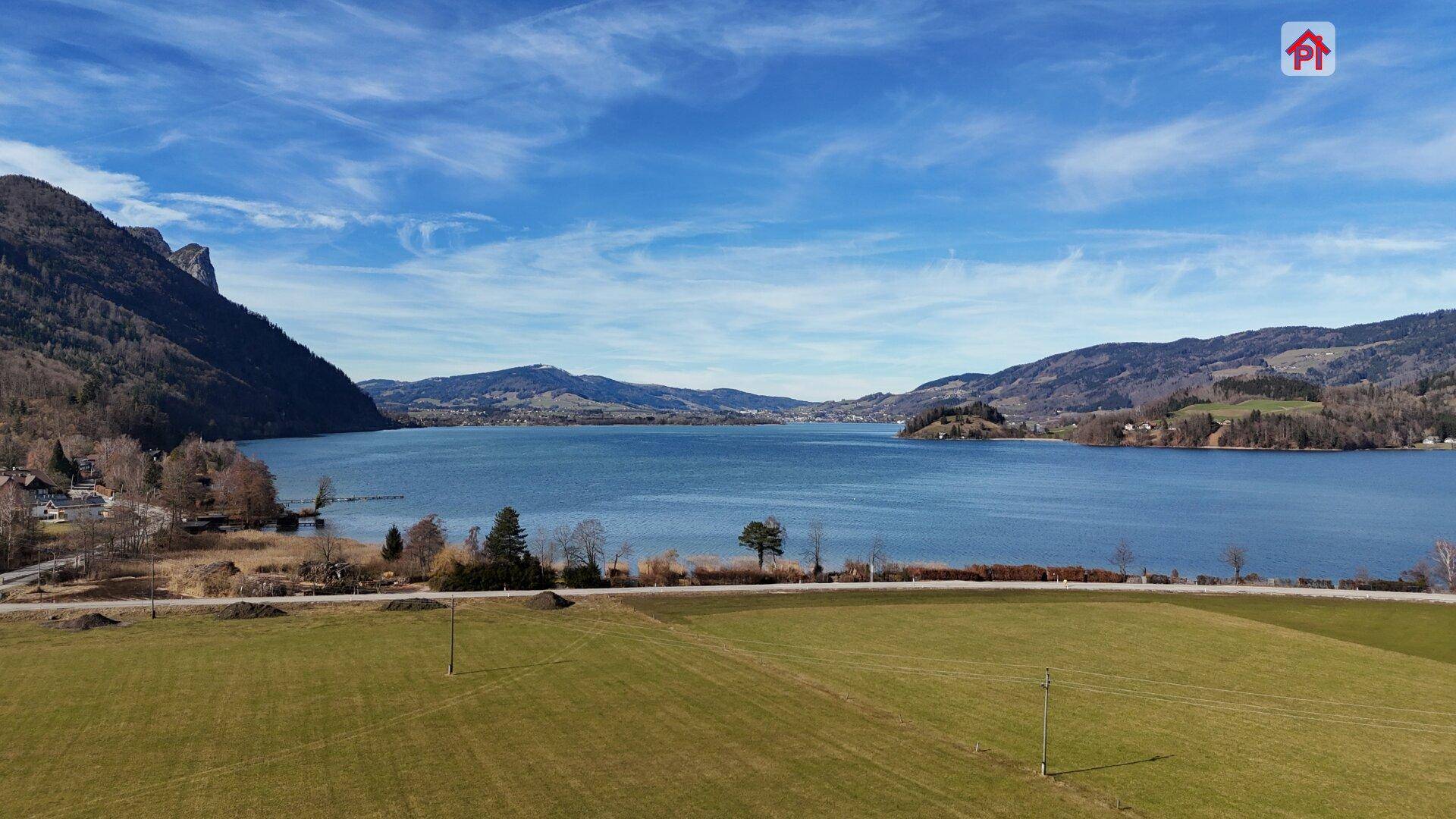 Weitläufige Landschaftsaufnahme mit einem großen See, umgeben von Bergen und grünen Feldern unter blauem Himmel.