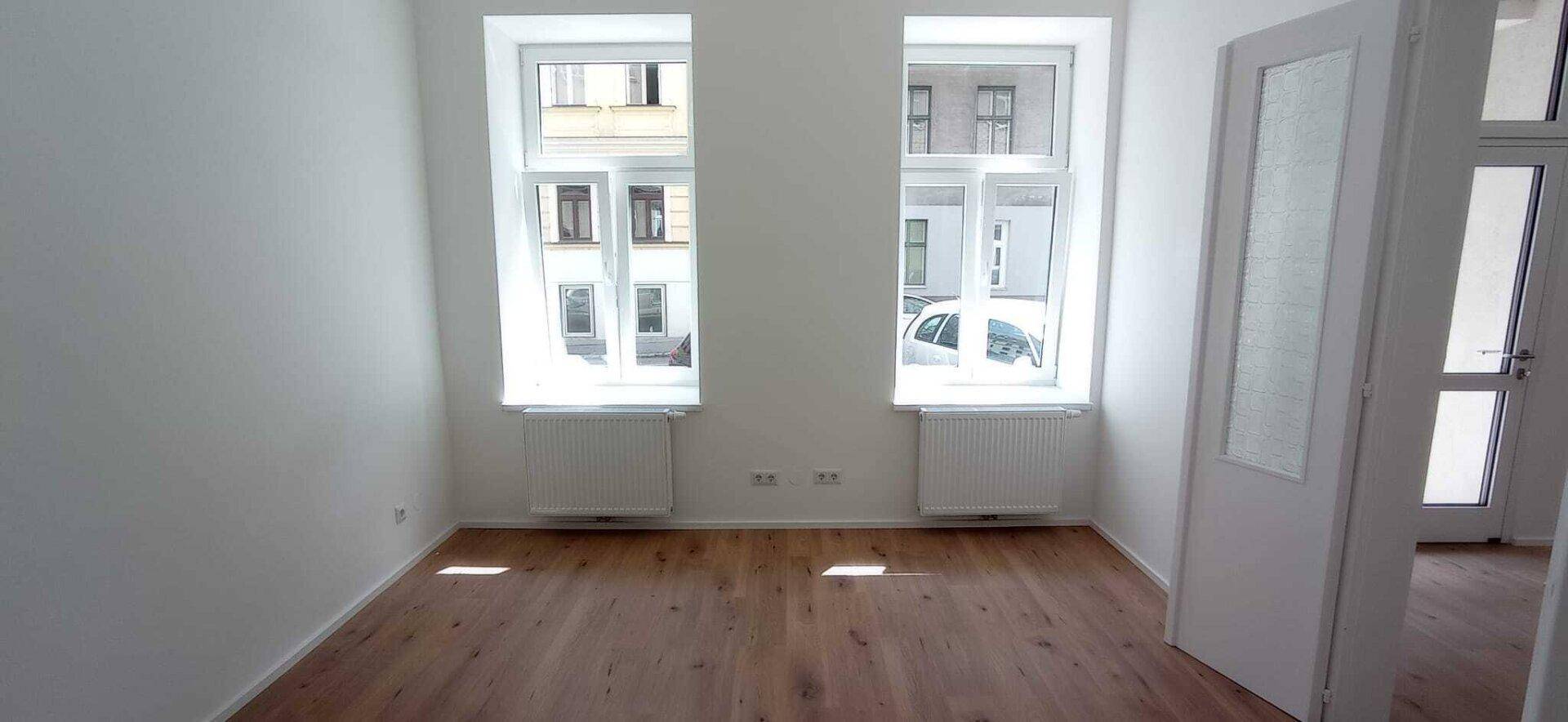 Heller, leerer Raum mit Holzboden, weißen Wänden und zwei großen Fenstern, die viel Licht spenden.