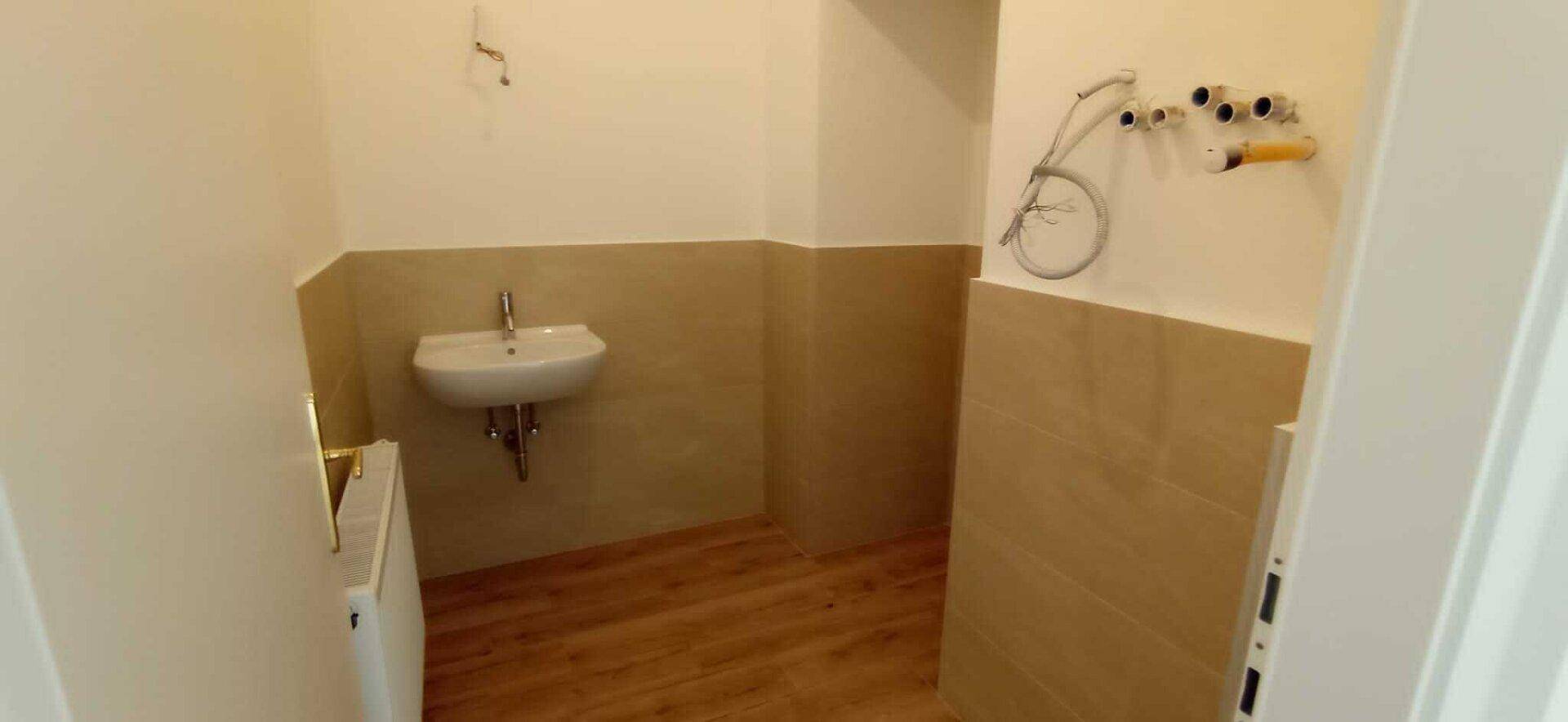 Badezimmer im Renovierungszustand mit Holzboden, beigefarbenen Fliesen, Waschbecken und Duschanschlüssen.