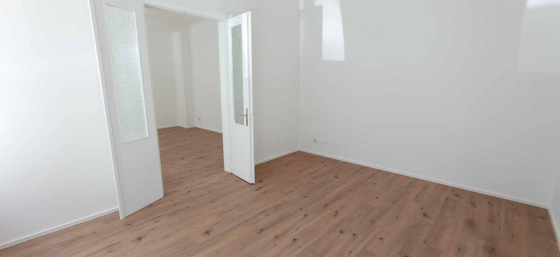 Geräumiger Raum mit Holzboden und weißen Wänden, verbunden durch eine Doppeltür mit Milchglas.