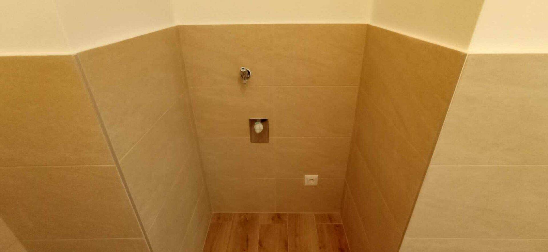 Badezimmerwand mit beigefarbenen Fliesen und Anschlüssen für eine Toilette, auf einem Holzboden.