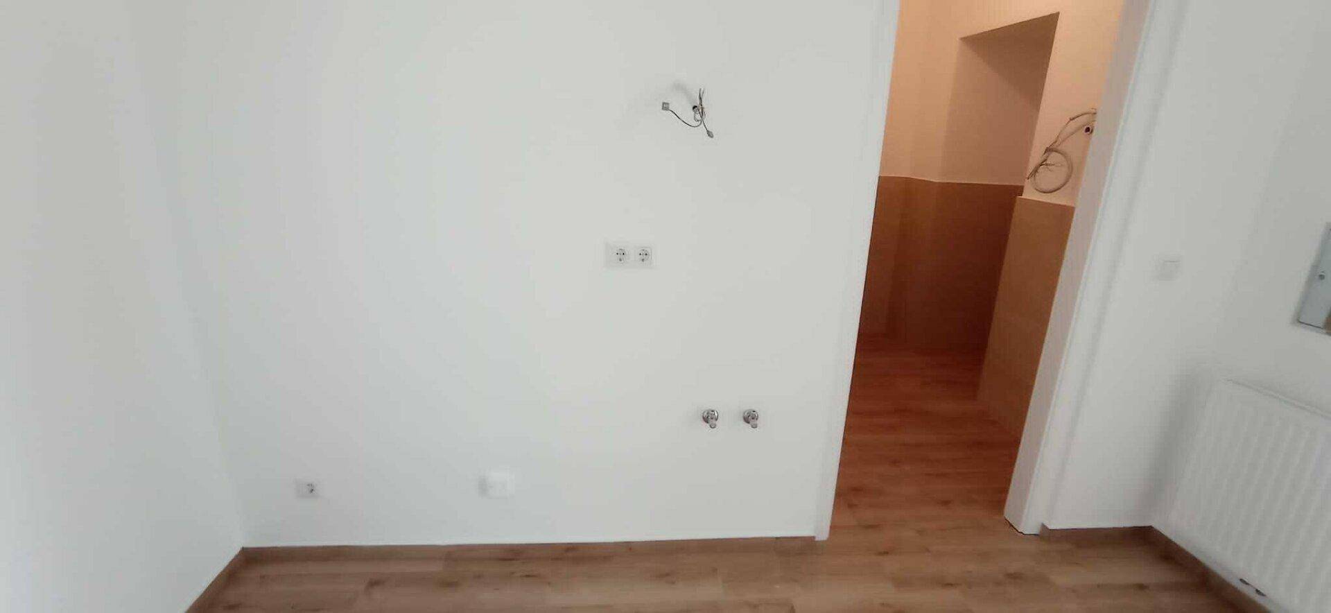 Raum mit Holzboden und weißen Wänden, sichtbaren Elektro- und Sanitäranschlüssen, angrenzend an einen gefliesten Bereich.