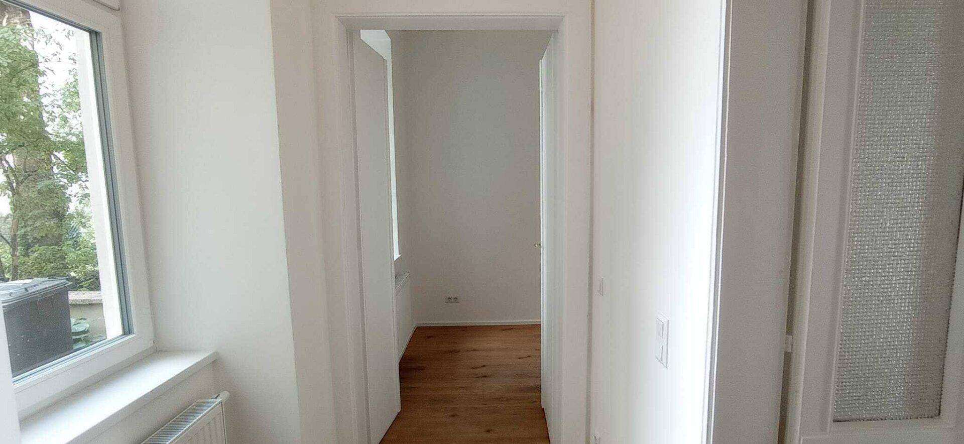 Heller Flur mit Holzboden und weißen Wänden, der zu mehreren Zimmern führt und ein Fenster am Ende hat.