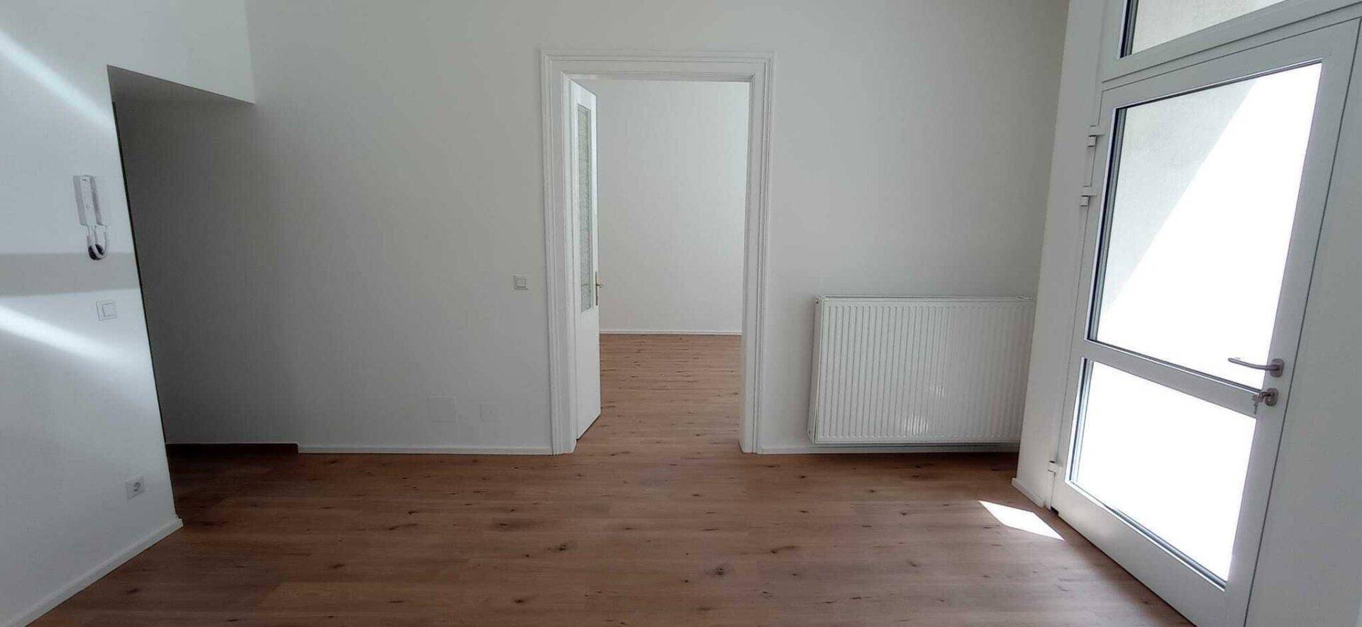 Heller Raum mit Holzboden, weißen Wänden und einem großen Fenster, das viel Tageslicht hereinlässt.