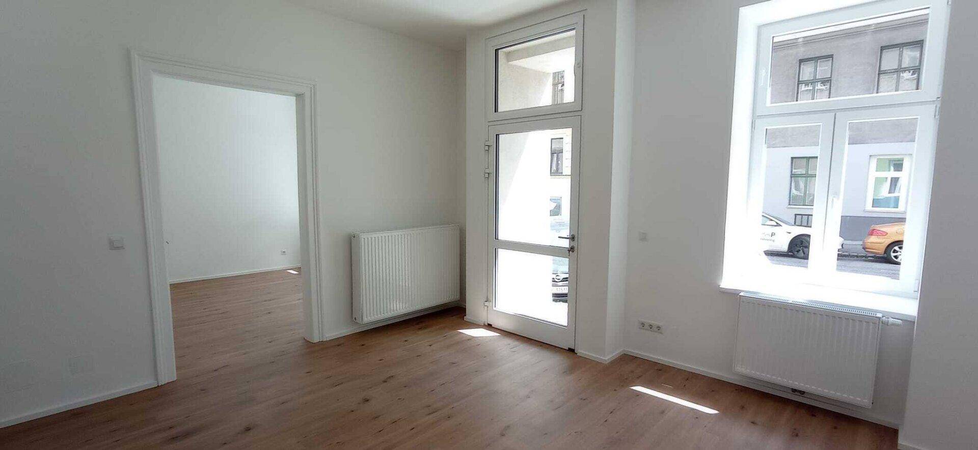 Geräumiger Wohnbereich mit Holzboden, großen Fenstern und einem Durchgang zu einem weiteren Zimmer.