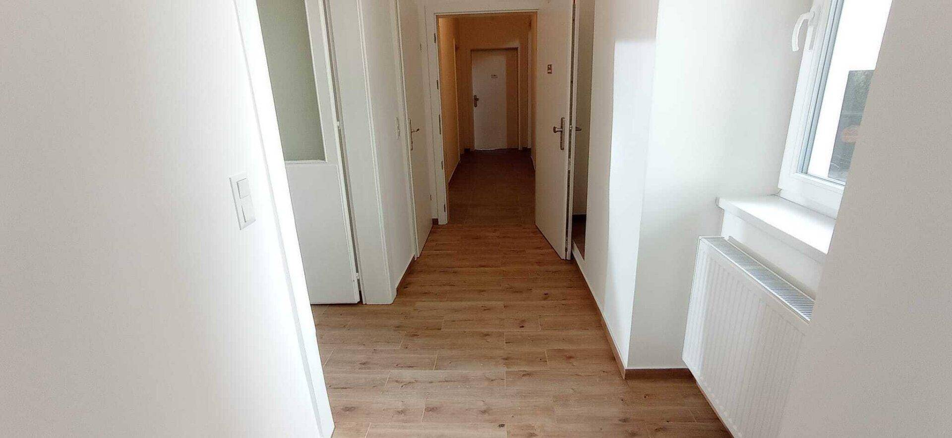 Langer Flur mit Holzboden und weißen Wänden, der zu verschiedenen Zimmern führt und am Ende ein Fenster hat.