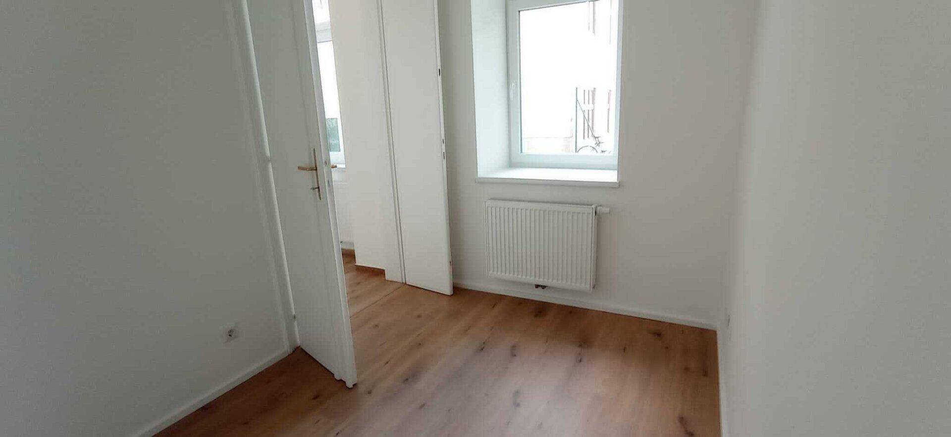 Helles Zimmer mit Holzboden, weißen Wänden, einem Fenster und einer offenen Tür, die den Raum zugänglich macht.
