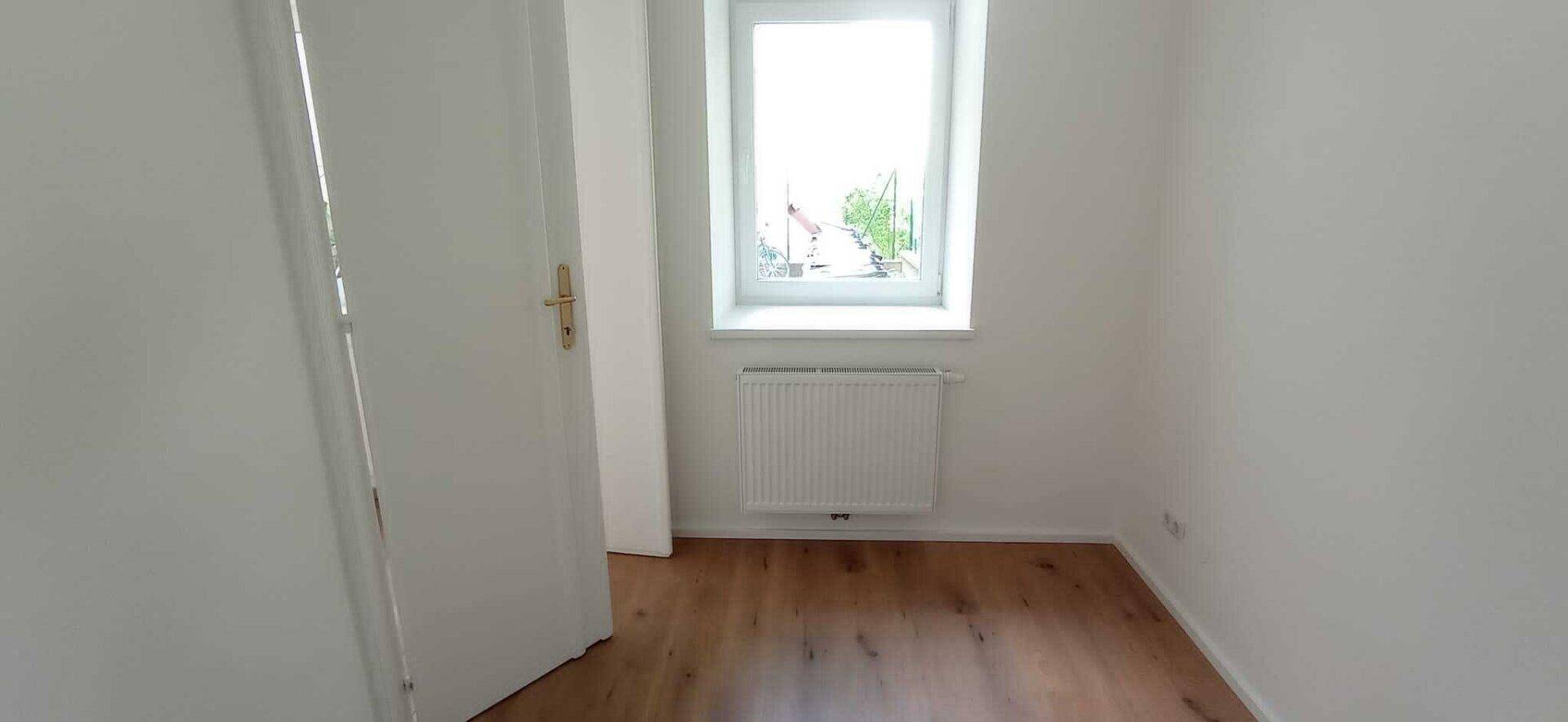 Gemütliches Zimmer mit Holzboden, weißen Wänden und einem Fenster, das den Raum mit Tageslicht füllt.