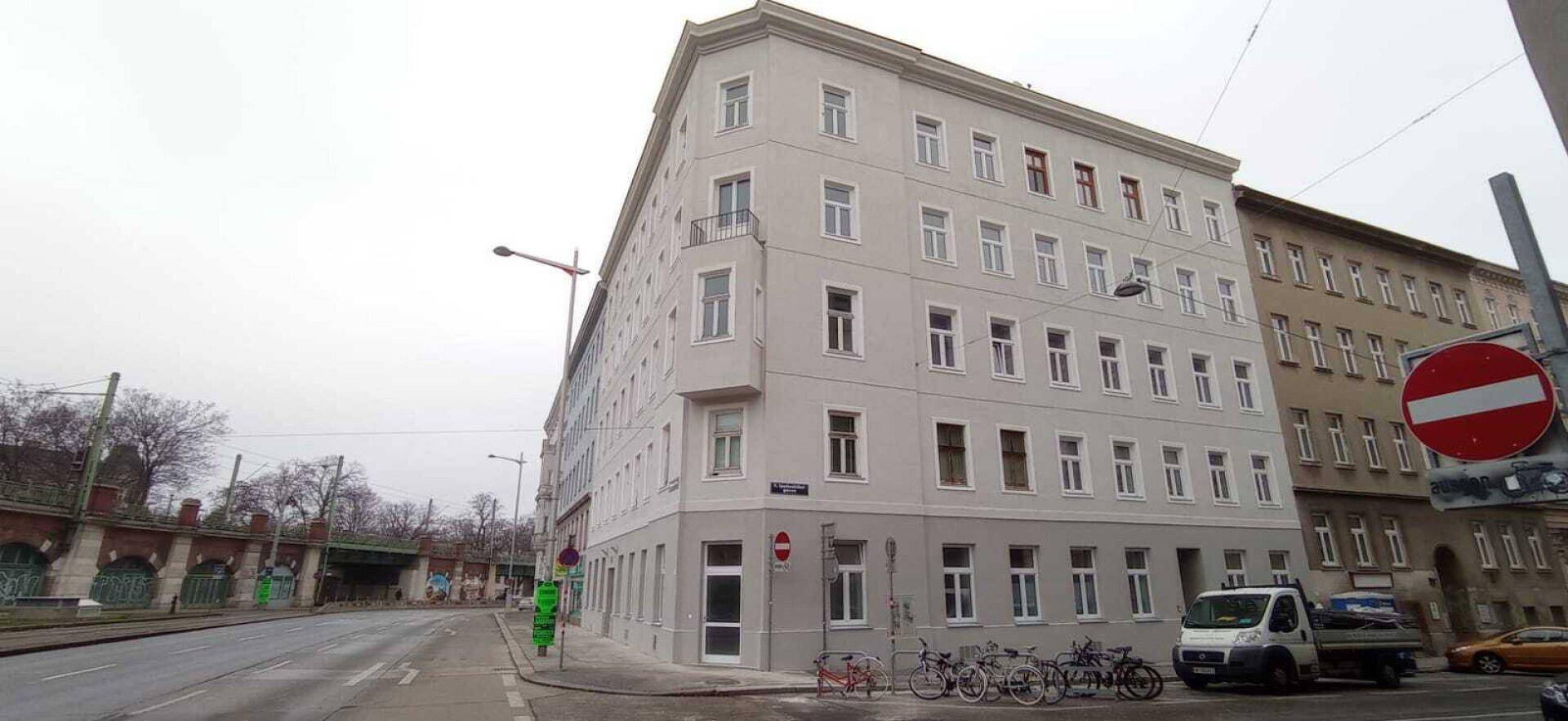Mehrstöckiges Eckgebäude an einer Stadtstraße unter bewölktem Himmel, mit Fahrrädern davor.