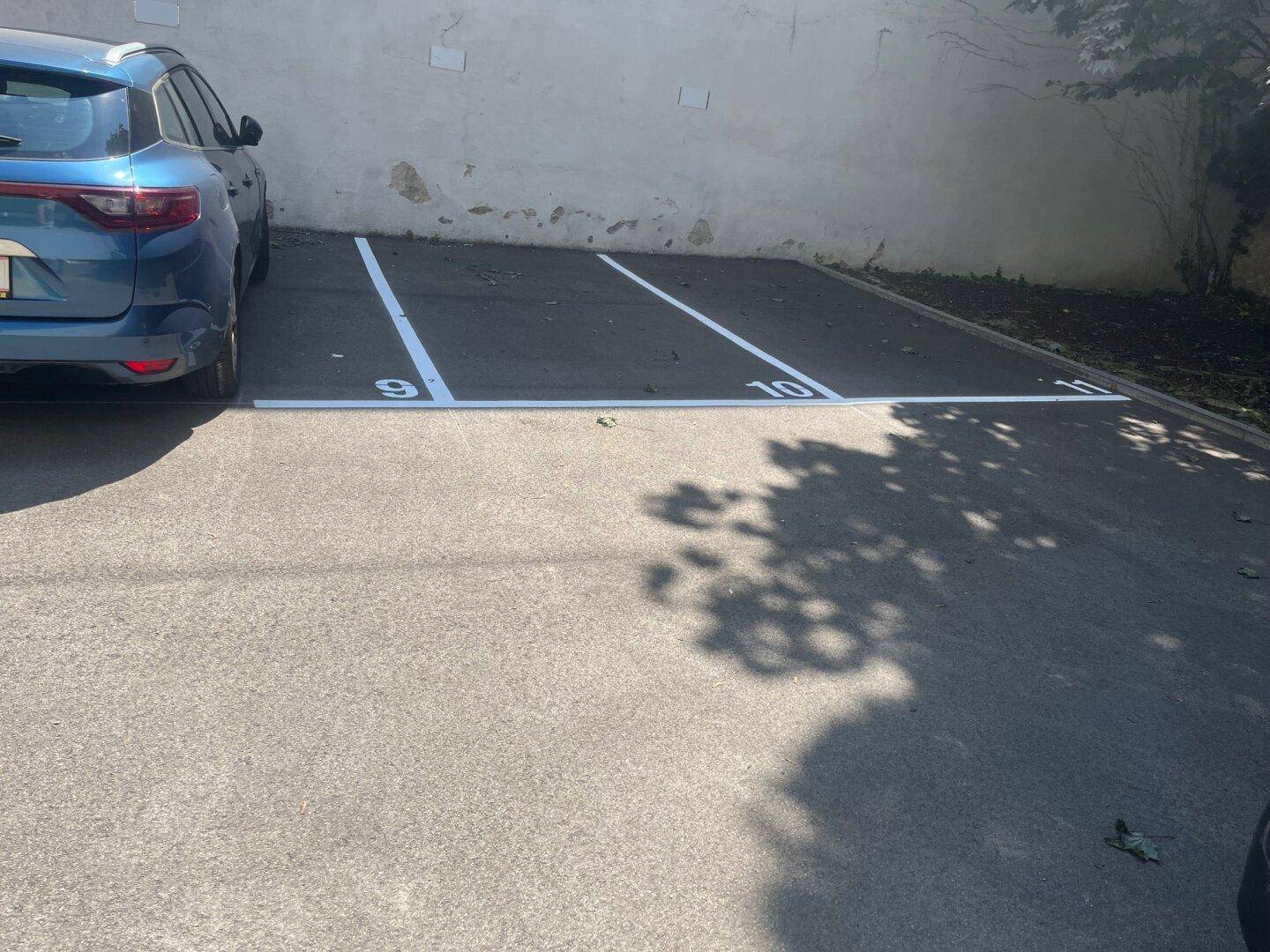 Markierte Außenparkplätze auf Asphalt neben einem blauen Auto und einer hellen Wand.