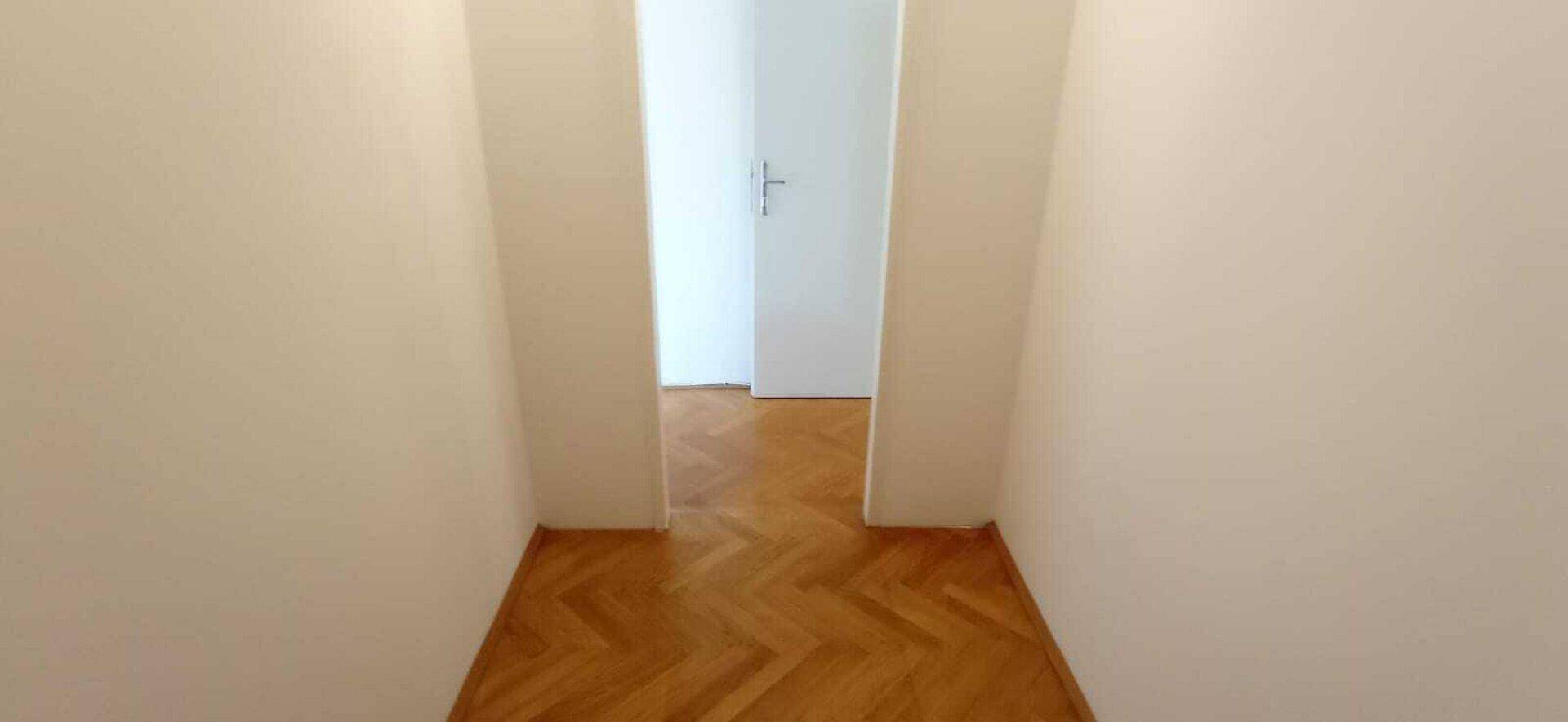 Schmaler Flur mit Fischgrätparkettboden und weißen Wänden, der zu weiteren Räumen führt.