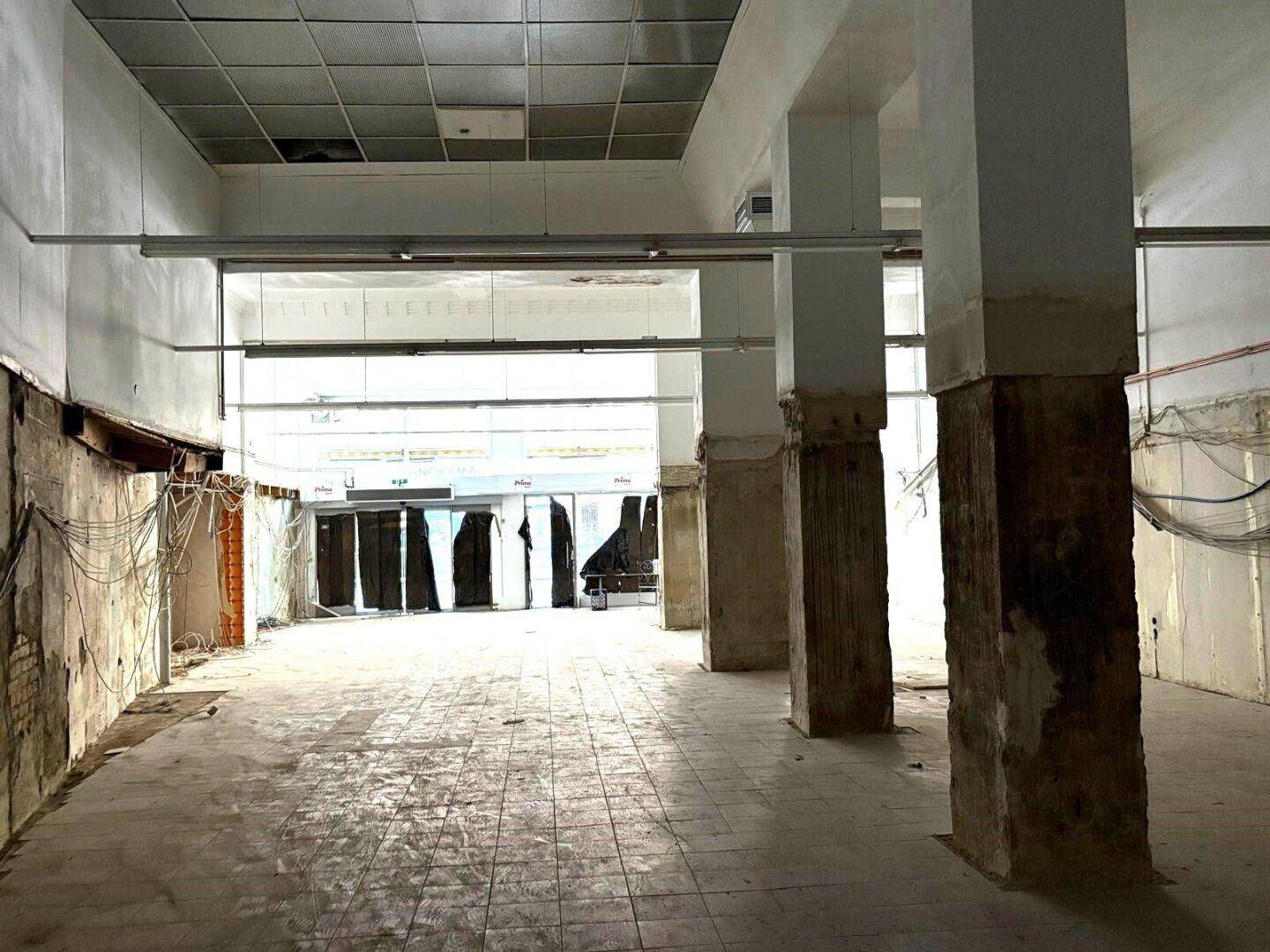 Weitläufiger Gewerberaum mit Säulen und großen Fenstern zur Straße, derzeit im Zustand der Entkernung.