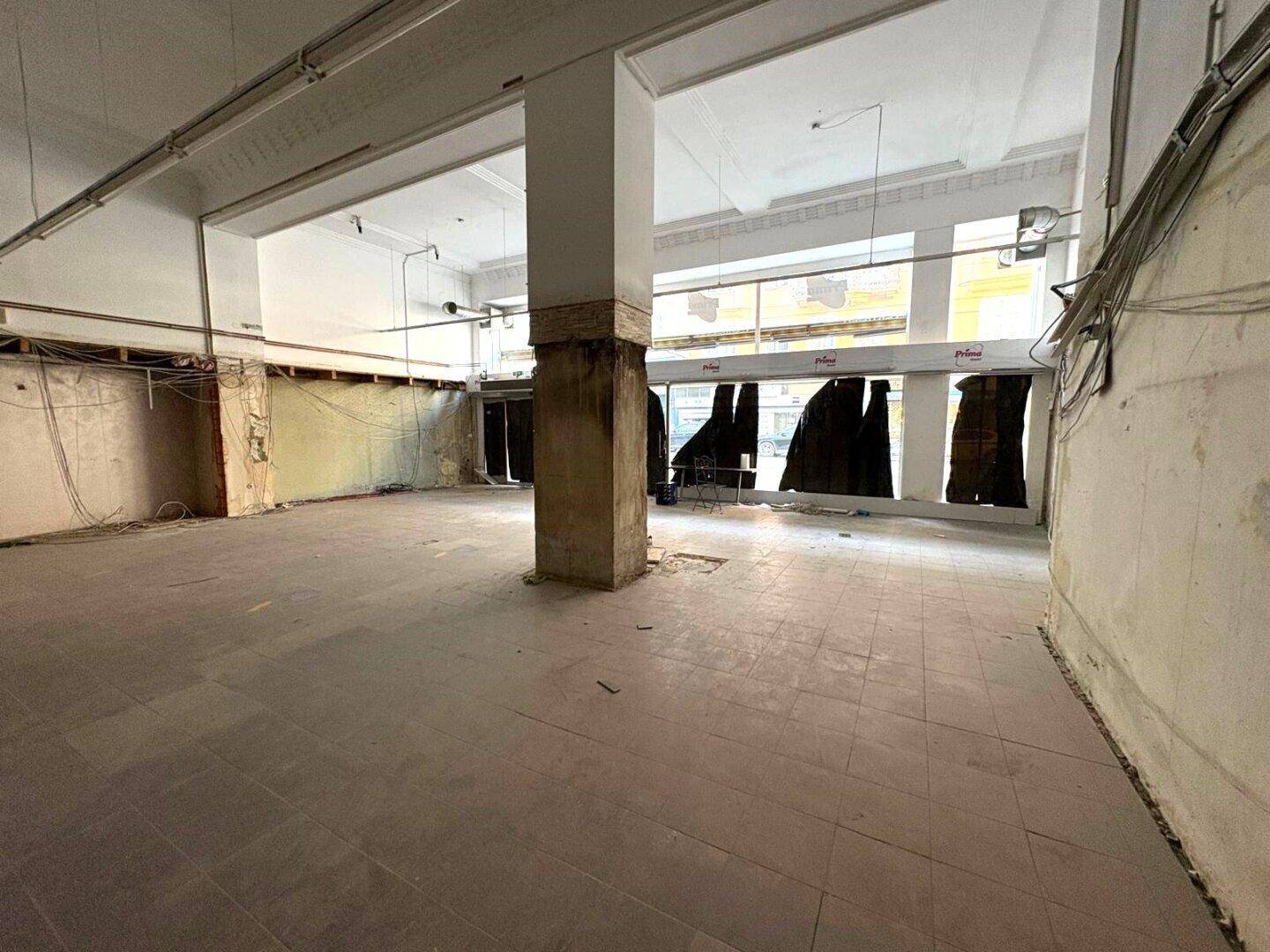 Weitläufiger Gewerberaum mit Säulen und großen Fenstern zur Straße, derzeit im Zustand der Entkernung.