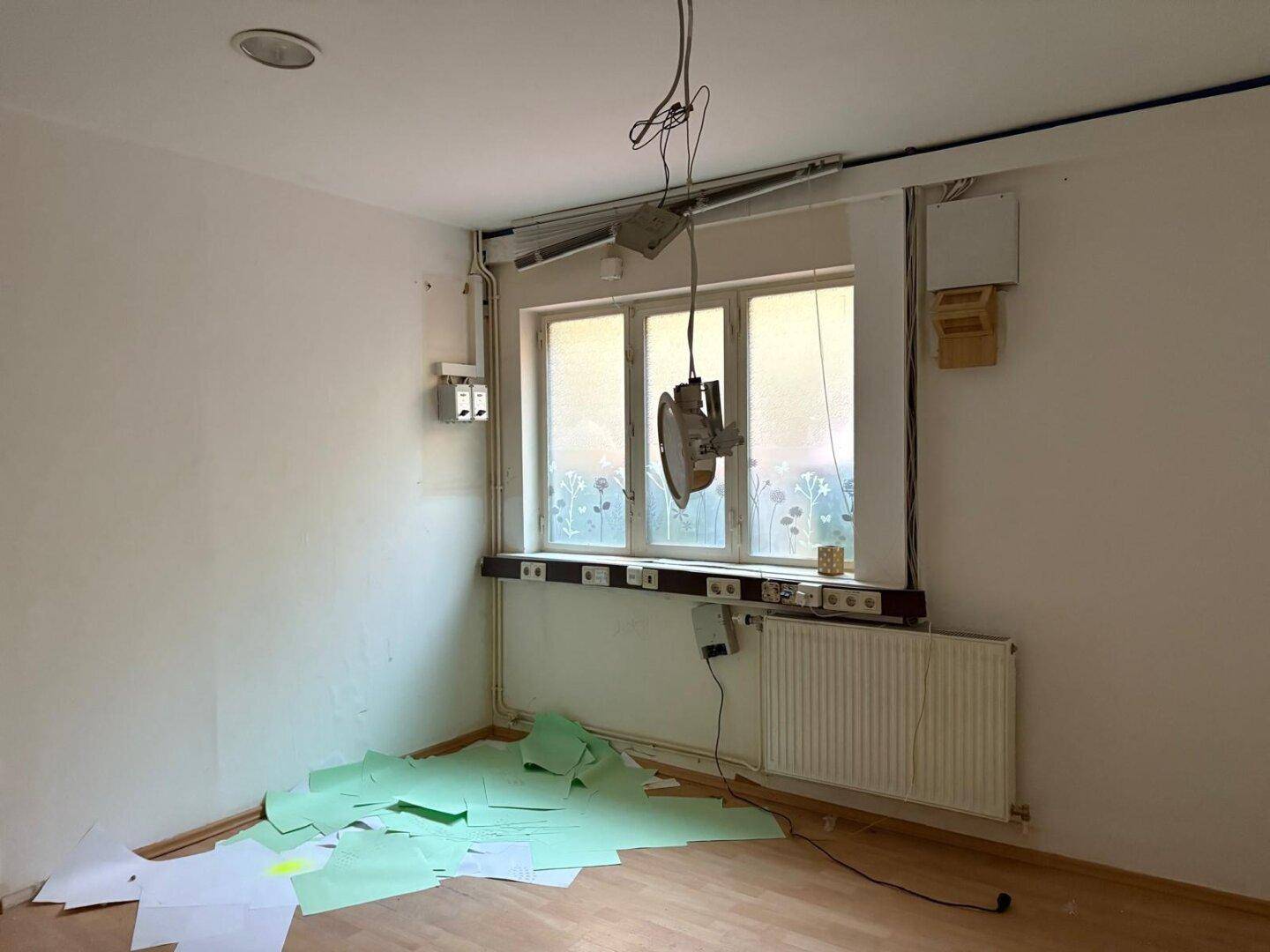 Heller Raum mit Holzfußboden und Fenster, ausgestattet mit Heizkörper und sichtbaren Elektroinstallationen.