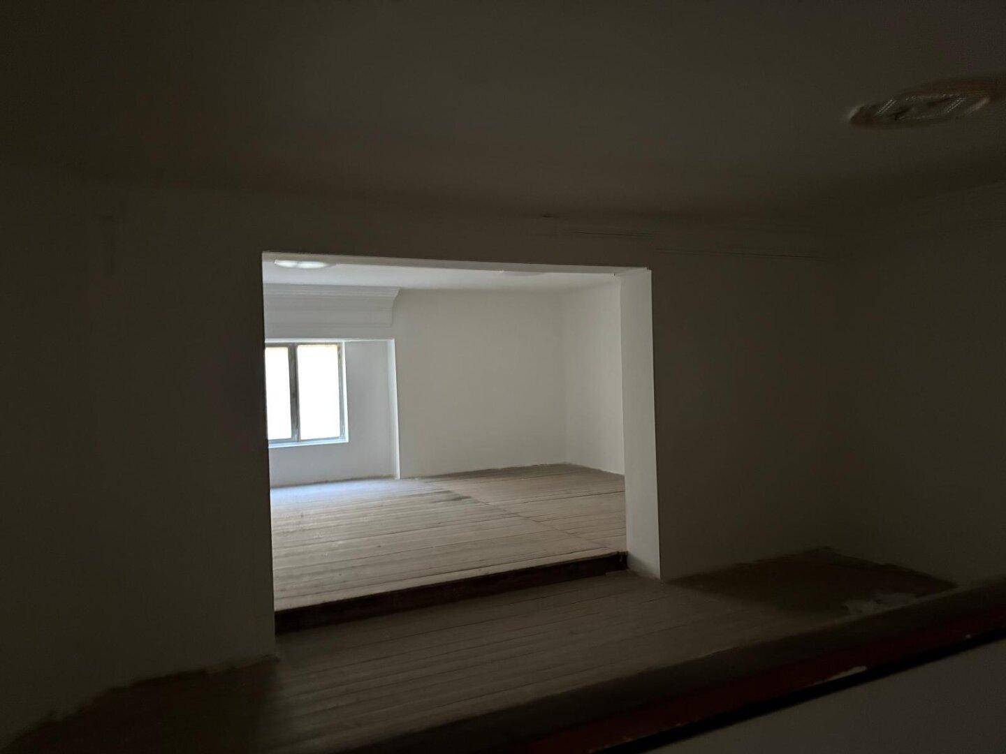 Blick durch einen Durchgang in einen helleren Raum mit Holzboden und Fenster, unmöbliert und leer.