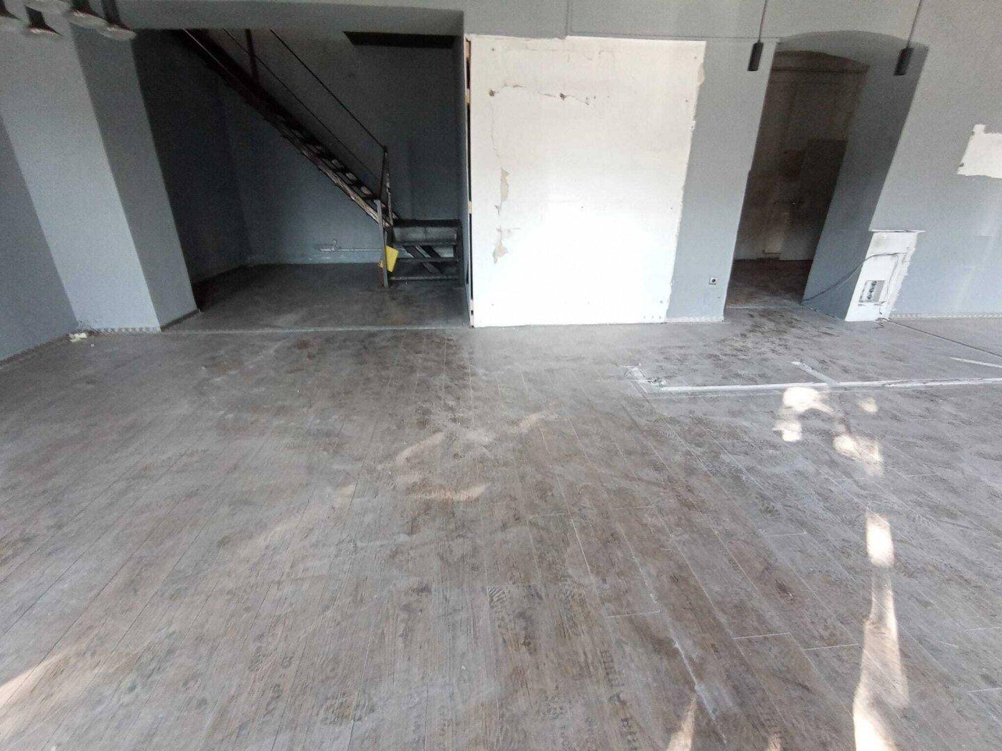 Innenansicht eines Geschäftsraums mit Holzboden, grauen Wänden und einer sichtbaren Treppe im Hintergrund.