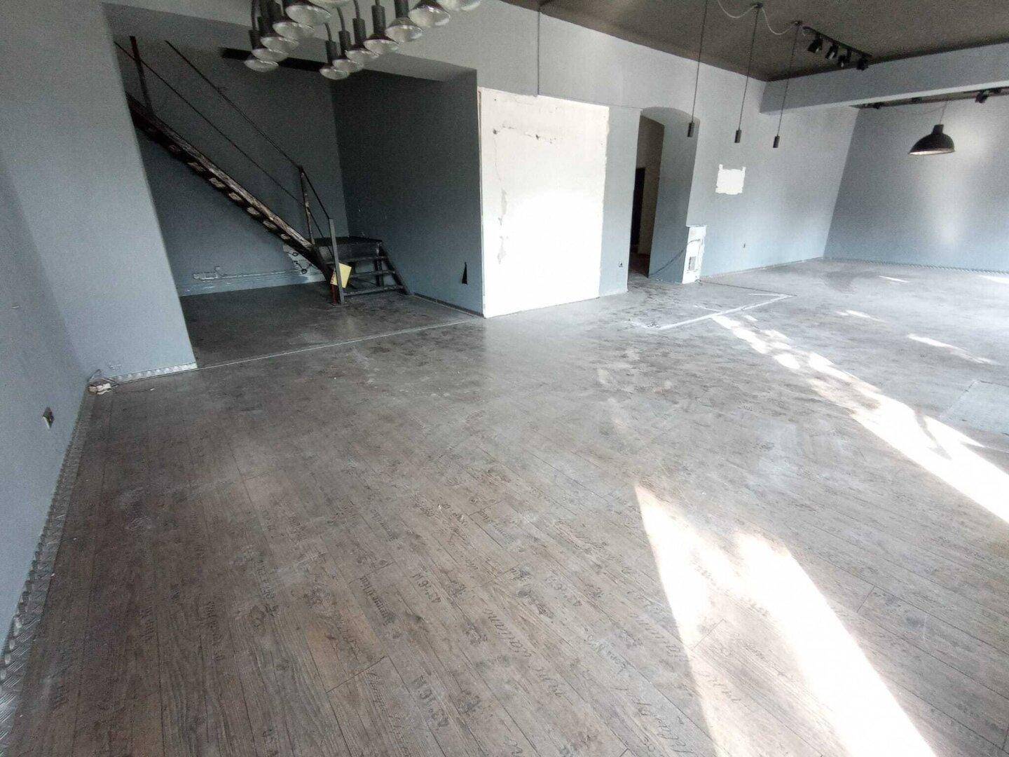 Geräumiger Geschäftsraum mit Holzboden, grauen Wänden und einer offenen Treppe zu einer oberen Ebene.