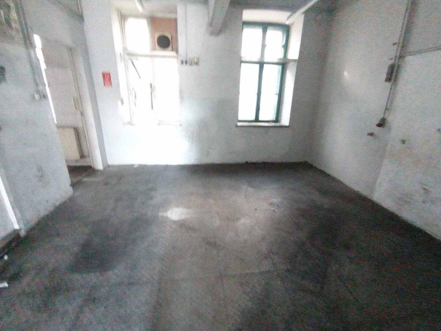 Heller Lagerraum mit Betonboden und weißen Wänden, der ein großes Fenster mit grünen Rahmen besitzt.