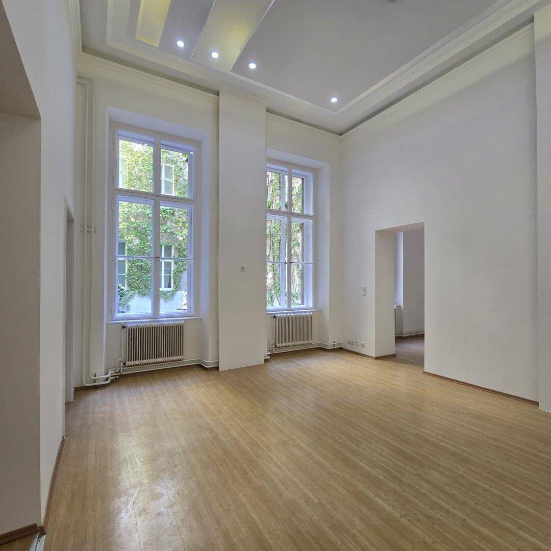 Heller Wohnraum mit hohen Decken, Parkettboden und Blick auf den grünen Innenhof durch große Fenster.