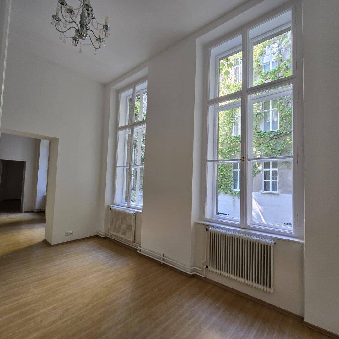 Geräumiges Wohnzimmer mit hohen Fenstern, Holzboden und einem eleganten Kronleuchter an der Decke.