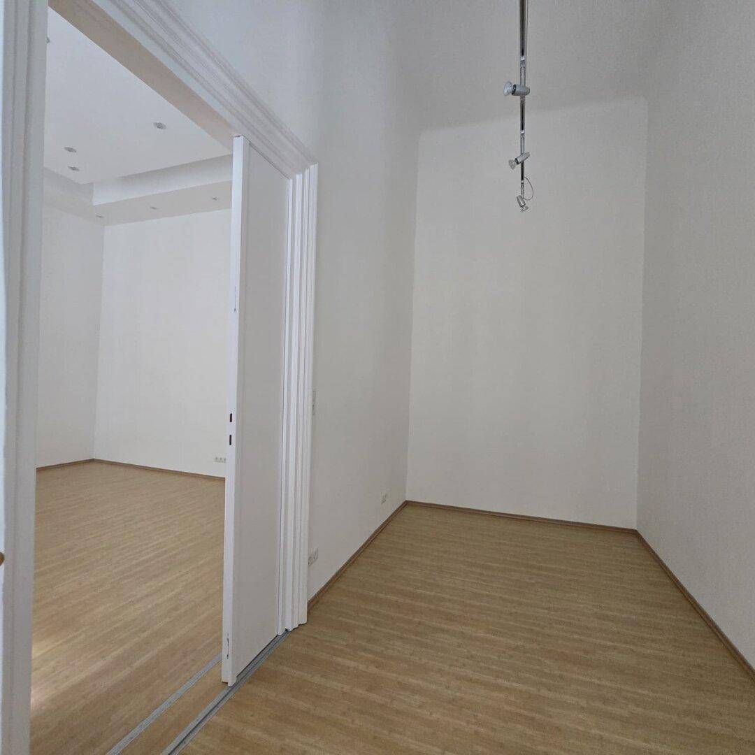 Heller Raum mit Holzboden und hoher Decke, angrenzend an einen weiteren Raum durch eine breite Öffnung.