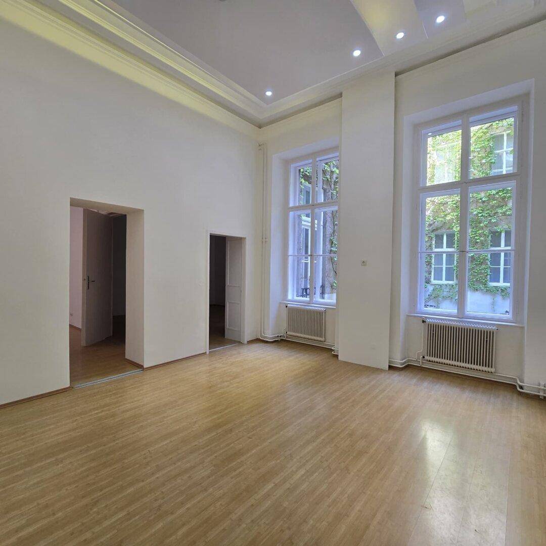 Geräumiges Wohnzimmer mit hohen Decken, Holzboden und großen Fenstern zum begrünten Innenhof.