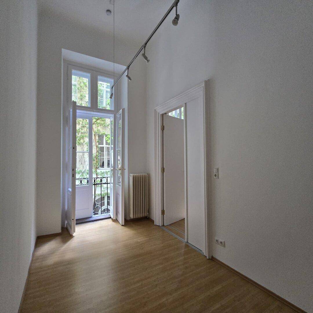 Langer, schmaler Raum mit Holzboden, hohen Decken und einem französischen Balkon zum Innenhof.