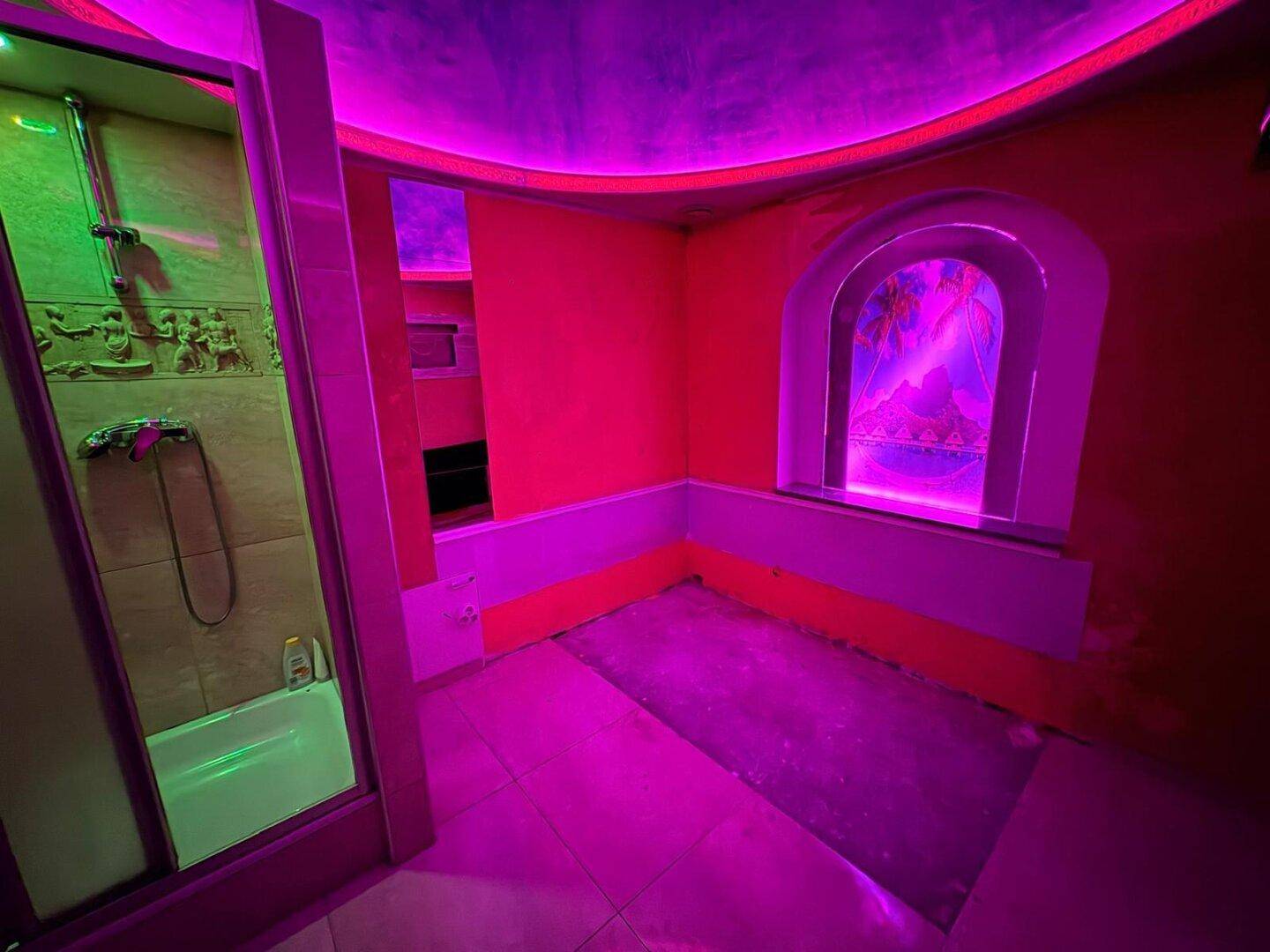 Raum mit pinker und roter Beleuchtung, Fliesenboden und einer Duschkabine, mit dekorativem Fenster.