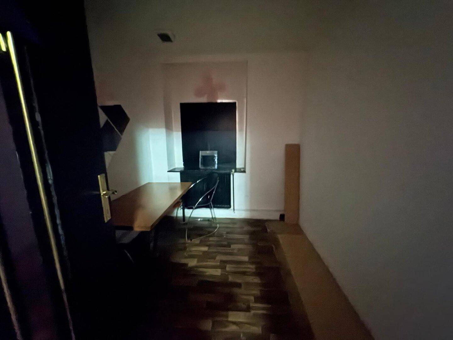 Schreibtischbereich in einem Raum mit Holzboden und weiß gestrichenen Wänden, teilweise dunkel.
