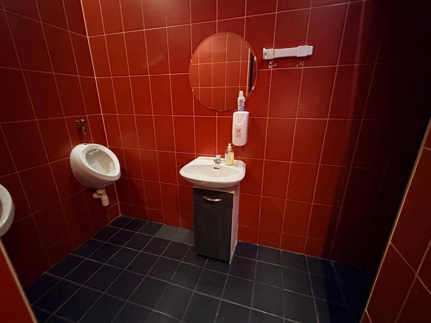 Badezimmer mit roten Fliesen, Urinal und Waschbecken mit Unterschrank und Spiegel.