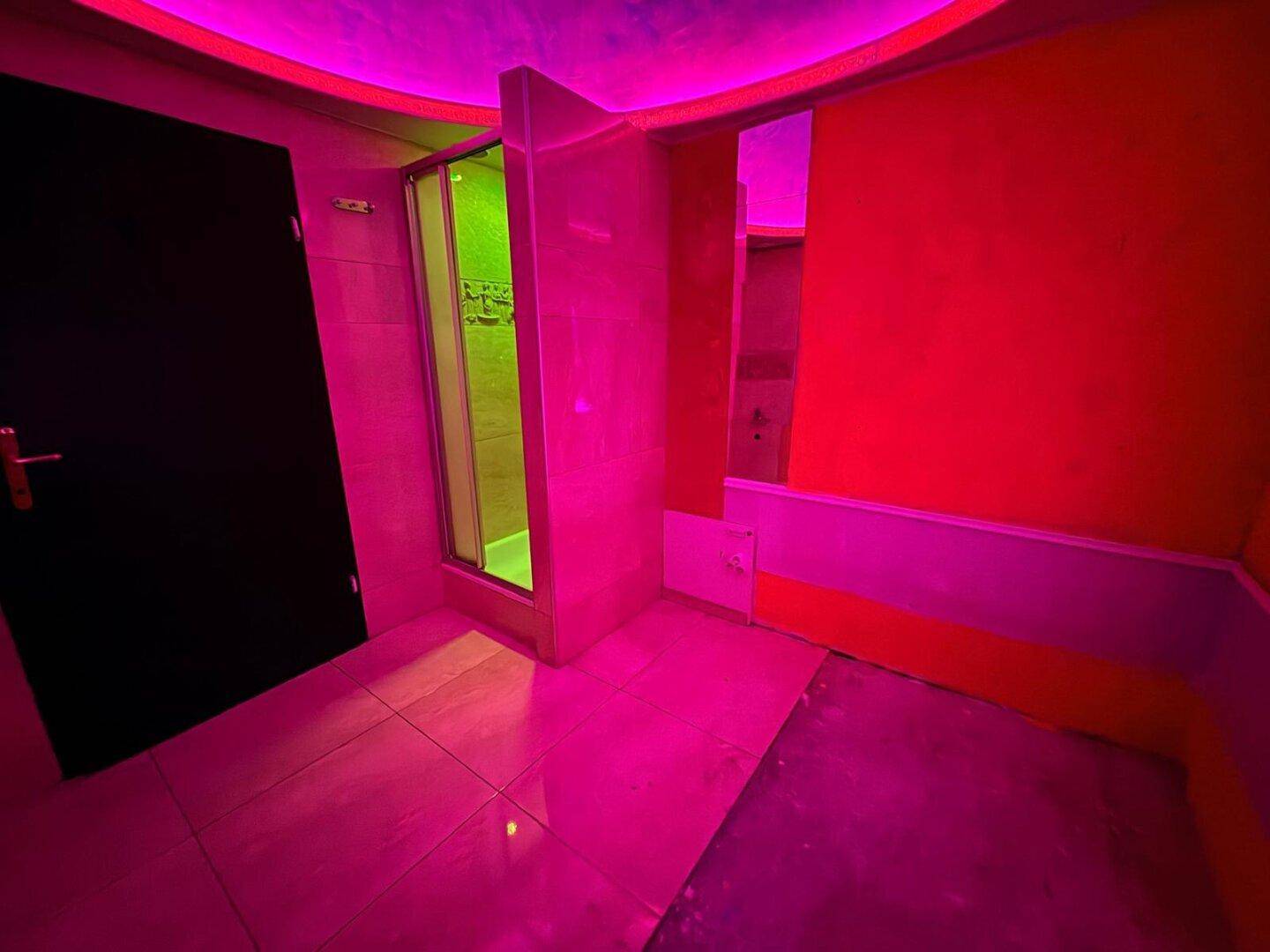 Raum mit pinker und roter Beleuchtung, Fliesenboden und einer Duschkabine, mit dunkler Tür.
