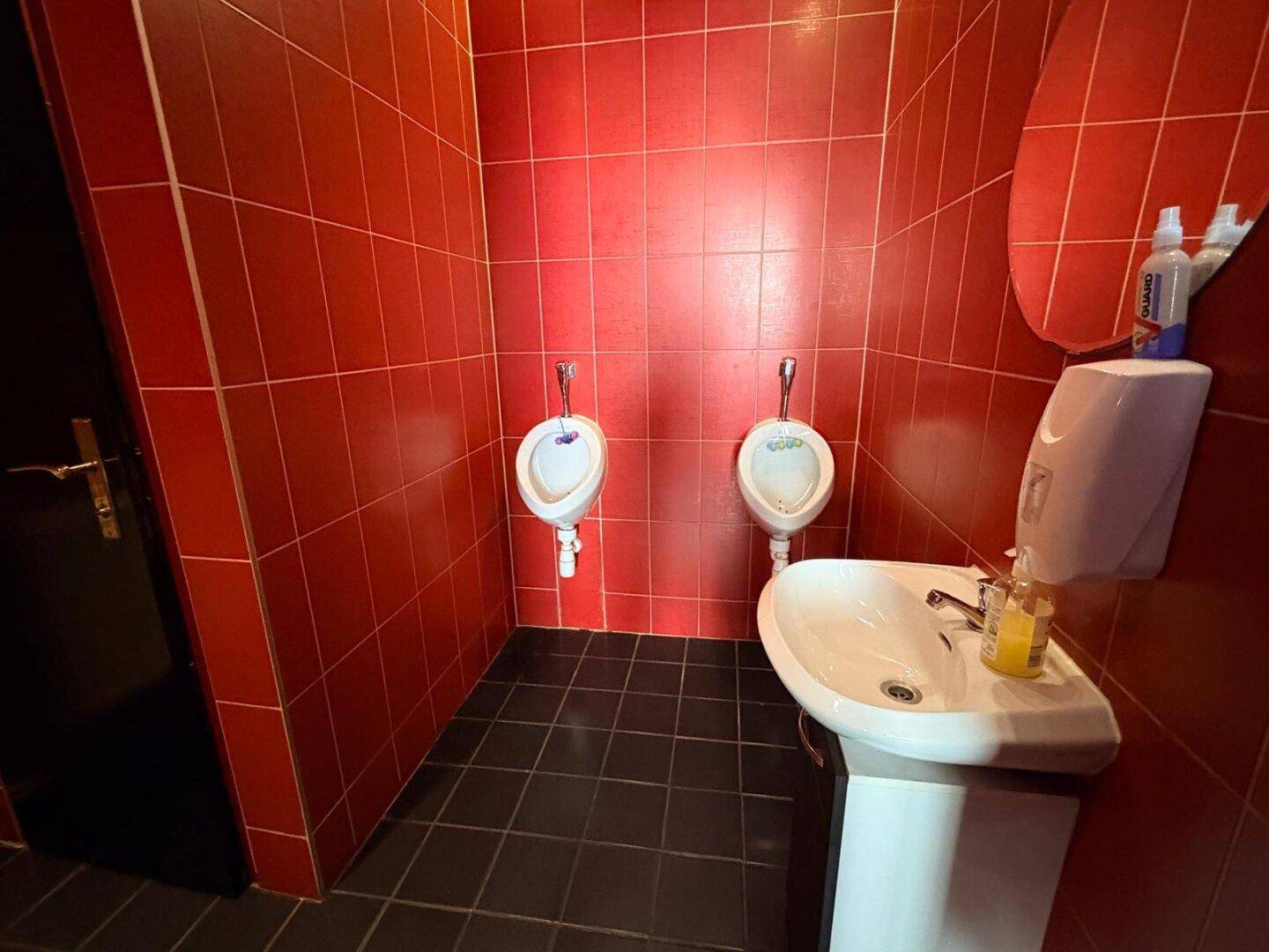 Badezimmer mit roten Fliesen, zwei Urinalen und einem Waschbecken mit Seifenspender.