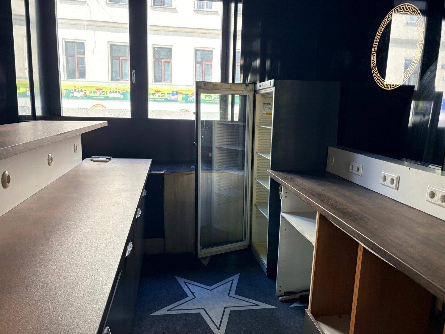 Thekenbereich mit Kühlschrank und Regalen, umgeben von großen Fenstern und Sternenmuster auf dem Boden.